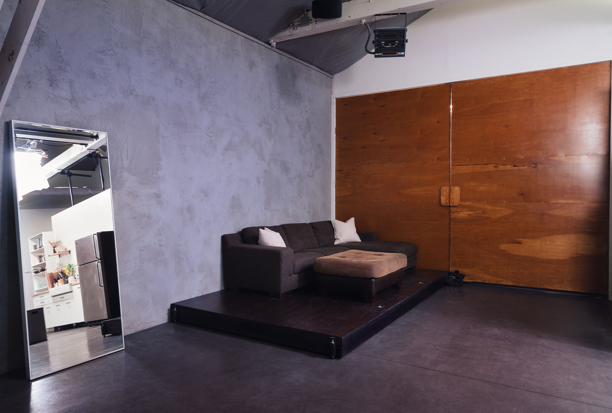 A couch on a stage on the studio floor of Particle Studio in Seattle, WA. A mirror in the corner reveals the kitchen that is off to the right side of camera, out of frame.