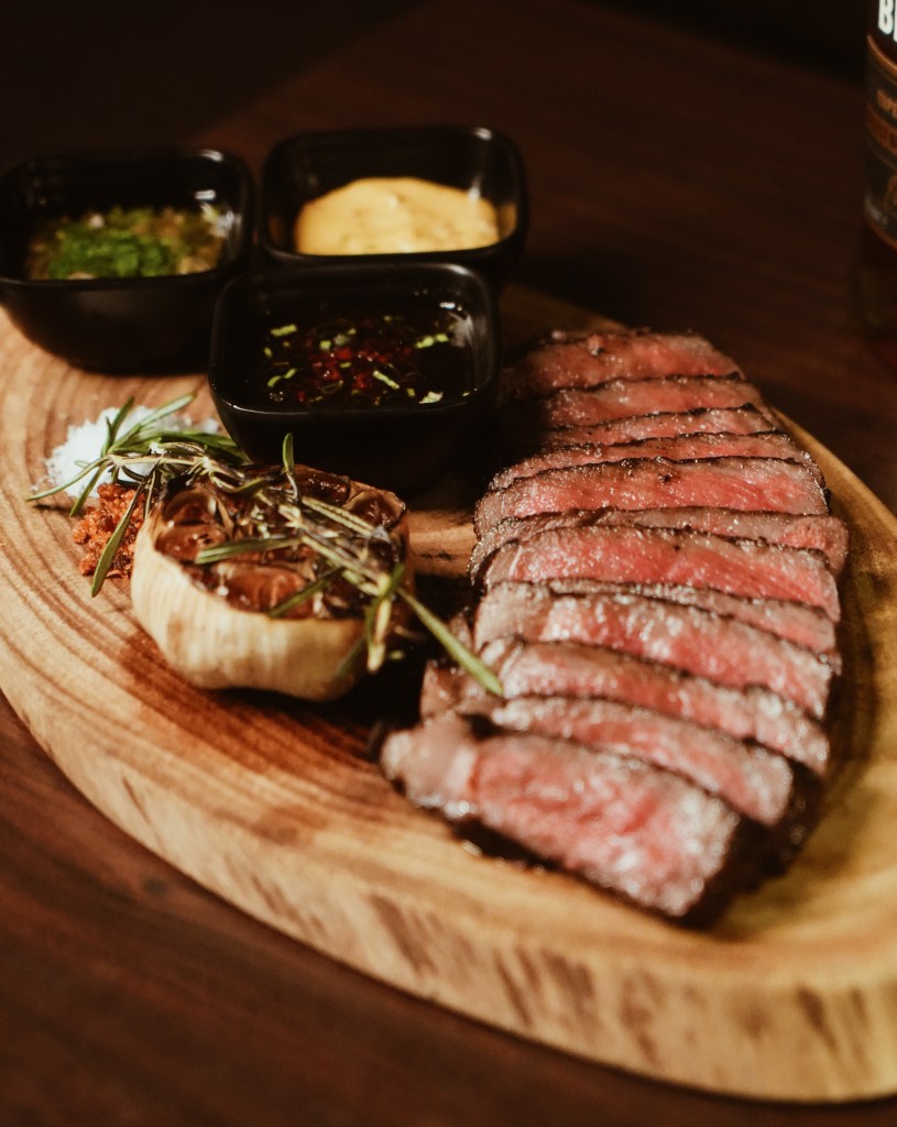 A beautifully arranged plate featuring slices of perfectly cooked meat, garnished with herbs, served on a wooden board alongside three small bowls of sauces.