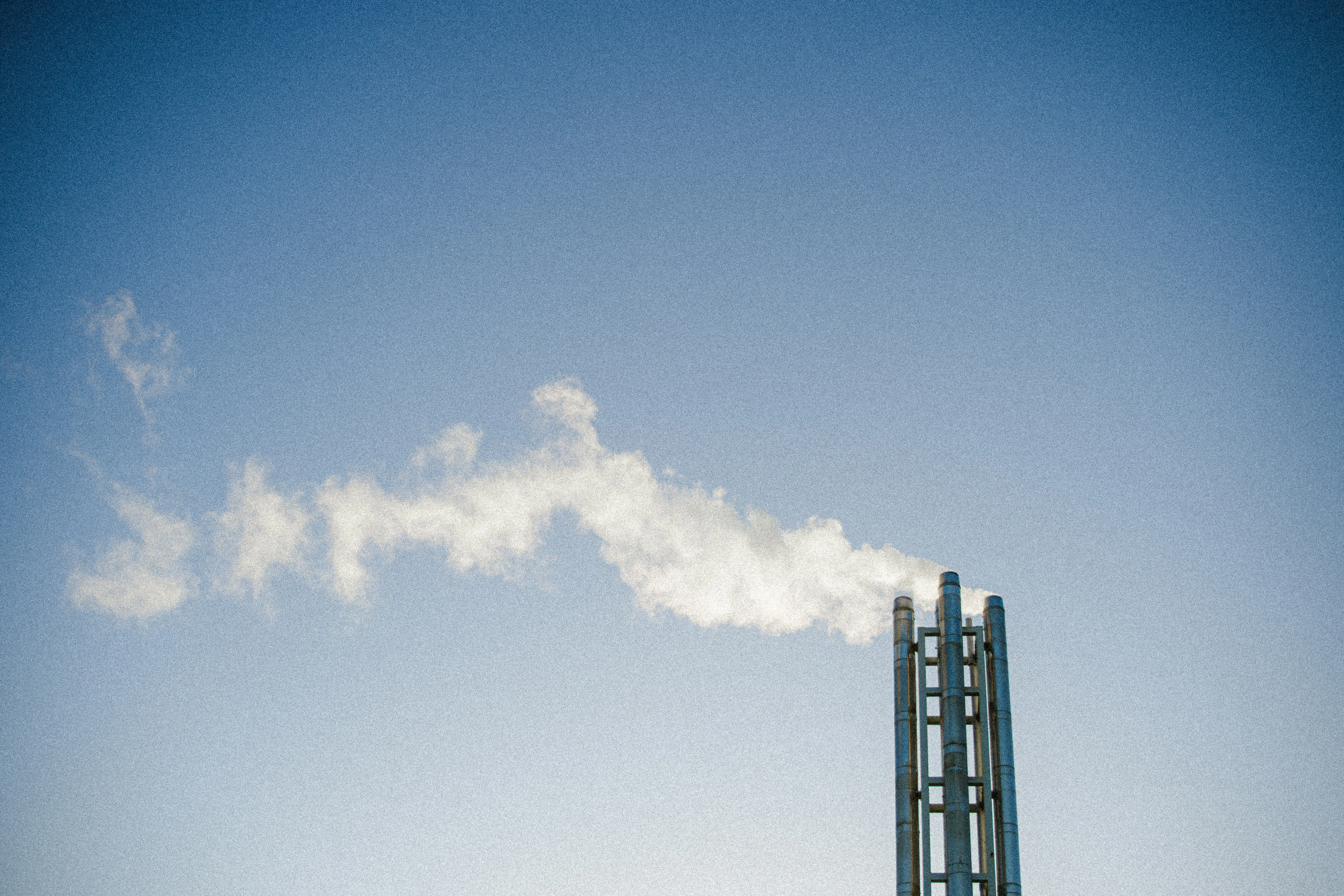 A tall industrial chimney stands against a clear blue sky, releasing a white plume that drifts away in the wind, illustrating Euroceptโs commitment to reaching zero emissions.