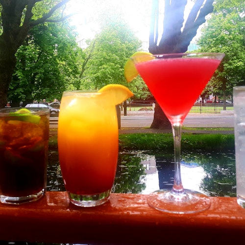 Four colorful cocktails, including a red martini, are displayed on a railing with a greenery-filled park in the background.
