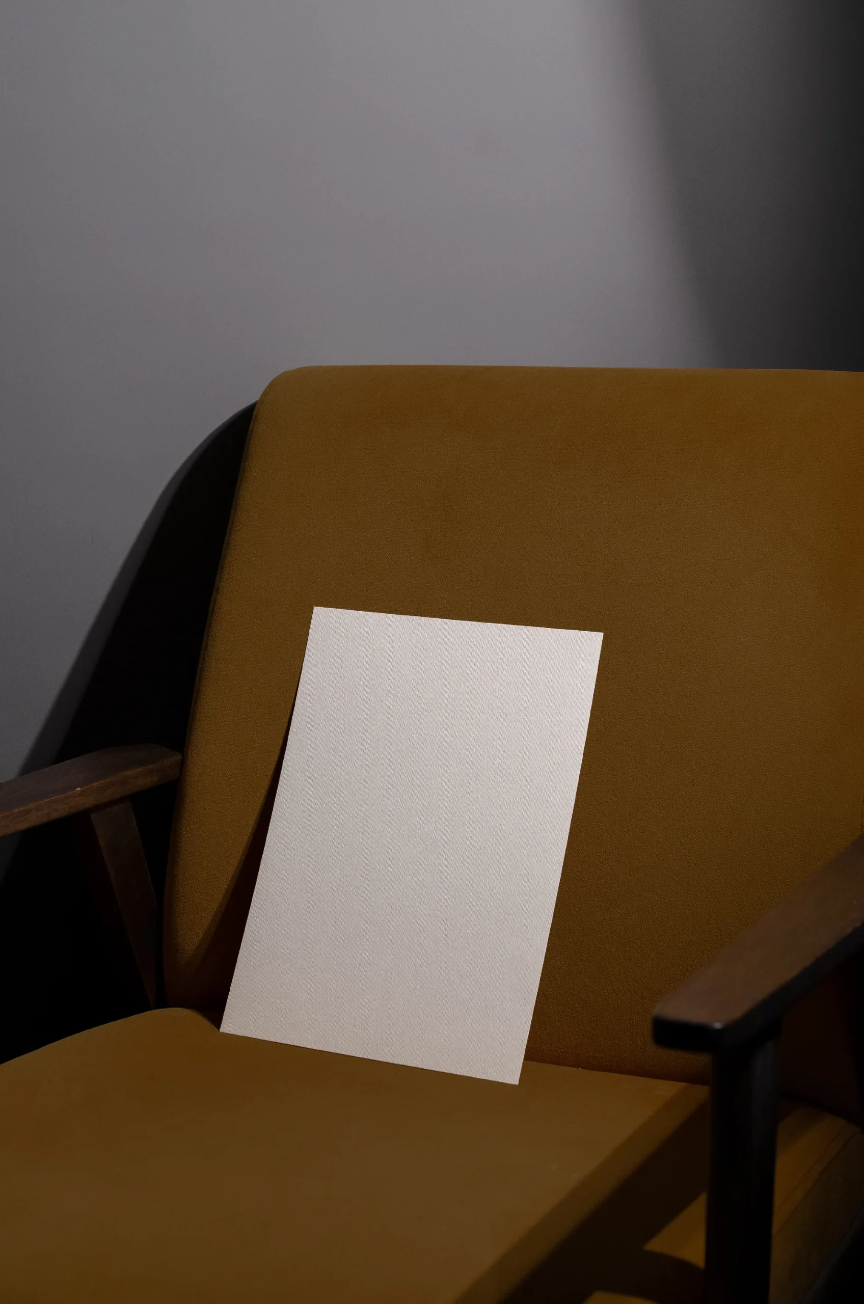 Minimalist mockup featuring a blank white textured paper placed on a mustard-yellow upholstered chair with wooden armrests, set against a neutral gray background with soft lighting.
