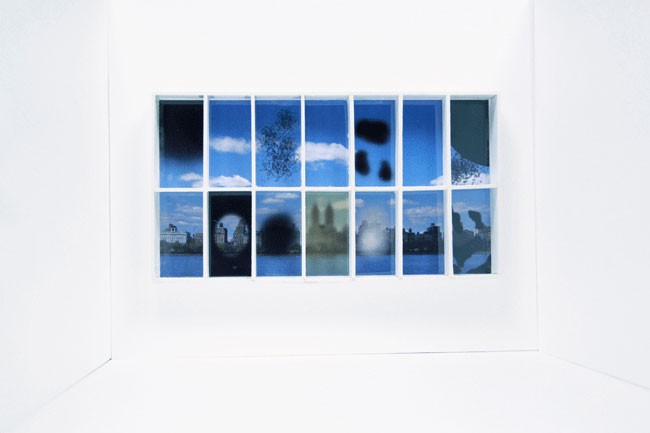 The image shows a white room with a white wall and a white baseboard. Mounted on the wall is a grid of 16 square photographs arranged in a 4x4 pattern. Each photograph depicts a blue-tinted scene: some show snowy landscapes, some show buildings, and some show abstract shapes. The photographs are evenly spaced, creating a structured and organized display. The lighting in the room is bright and even, likely from artificial sources.