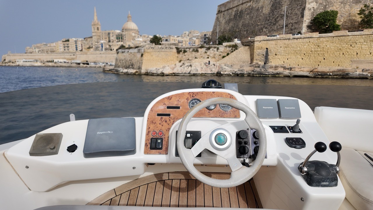 Sealine F37 flybridge helm station with burl wood dashboard, Raymarine chartplotters, and leather-wrapped steering wheel — open-air upper deck helm overlooking Valletta waterfront and St Paul's Cathedral dome, Malta