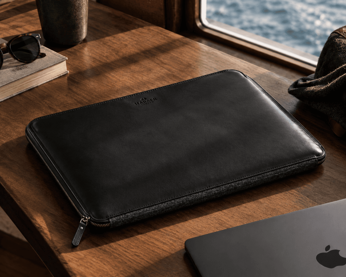 A premium tech accessory layout on a warm wooden desk inside a vessel cabin, with a porthole window revealing blue ocean waters outside. In the center remains a black leather laptop zip case with a dark gray felt lower panel and embossed Harber London branding. Beside the sleeve sits a dark gray laptop, a book topped with sunglasses, a dark green ceramic tumbler, and a folded jacket in the corner.