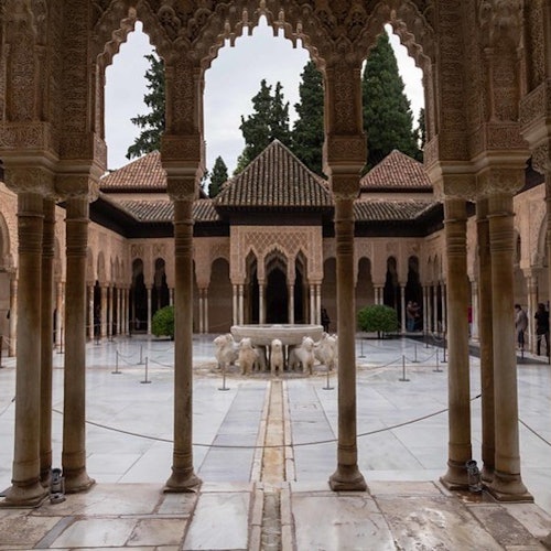 Patio con suelo de mármol, arcos de piedra decorativos, fuente central adornada con estatuas de leones y árboles al fondo.