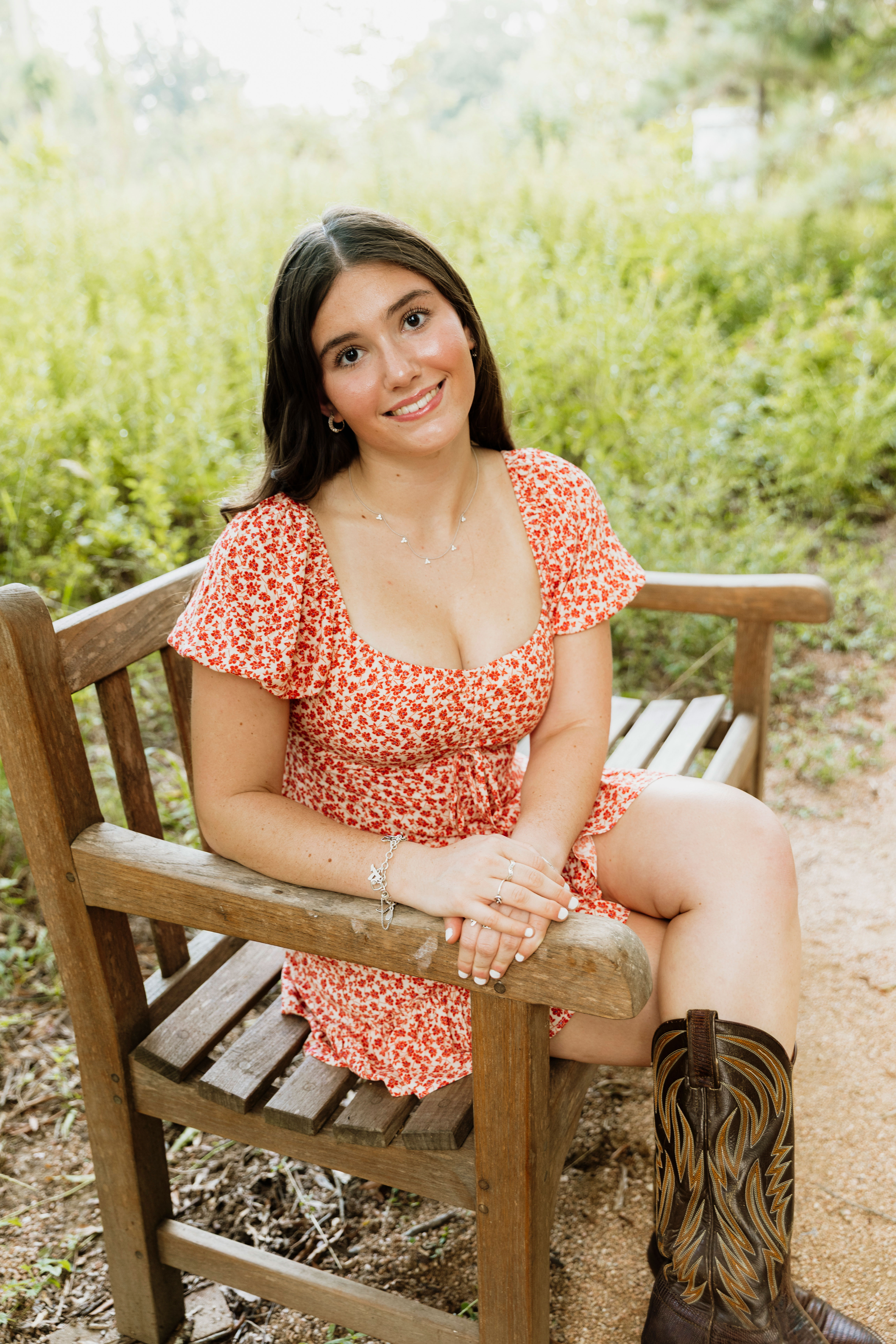 A high school senior girl sits smiling on a wooden bench at the Houston Arboretum. She is wearing a short, red and white floral dress paired with brown leather cowboy boots. Her long dark hair is parted down the middle, and she is wearing delicate silver jewelry. The background is a soft, out-of-focus lush green nature scene with bright, natural sunlight filtering through the trees.