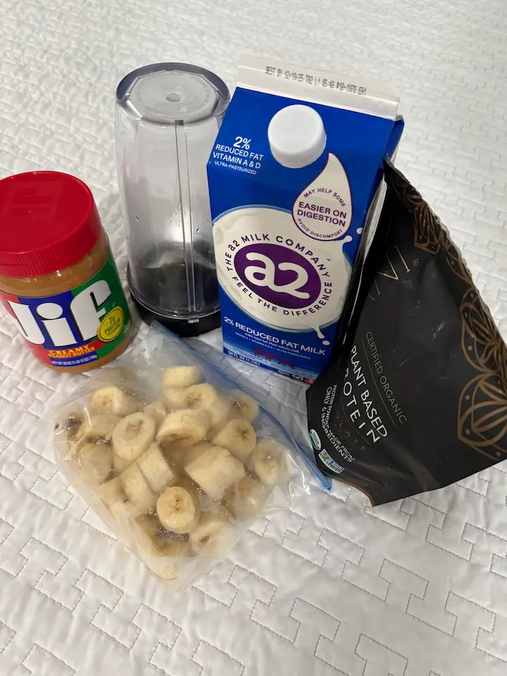 Chocolate whey protein, peanut butter, and frozen banana laid out beside a blender.