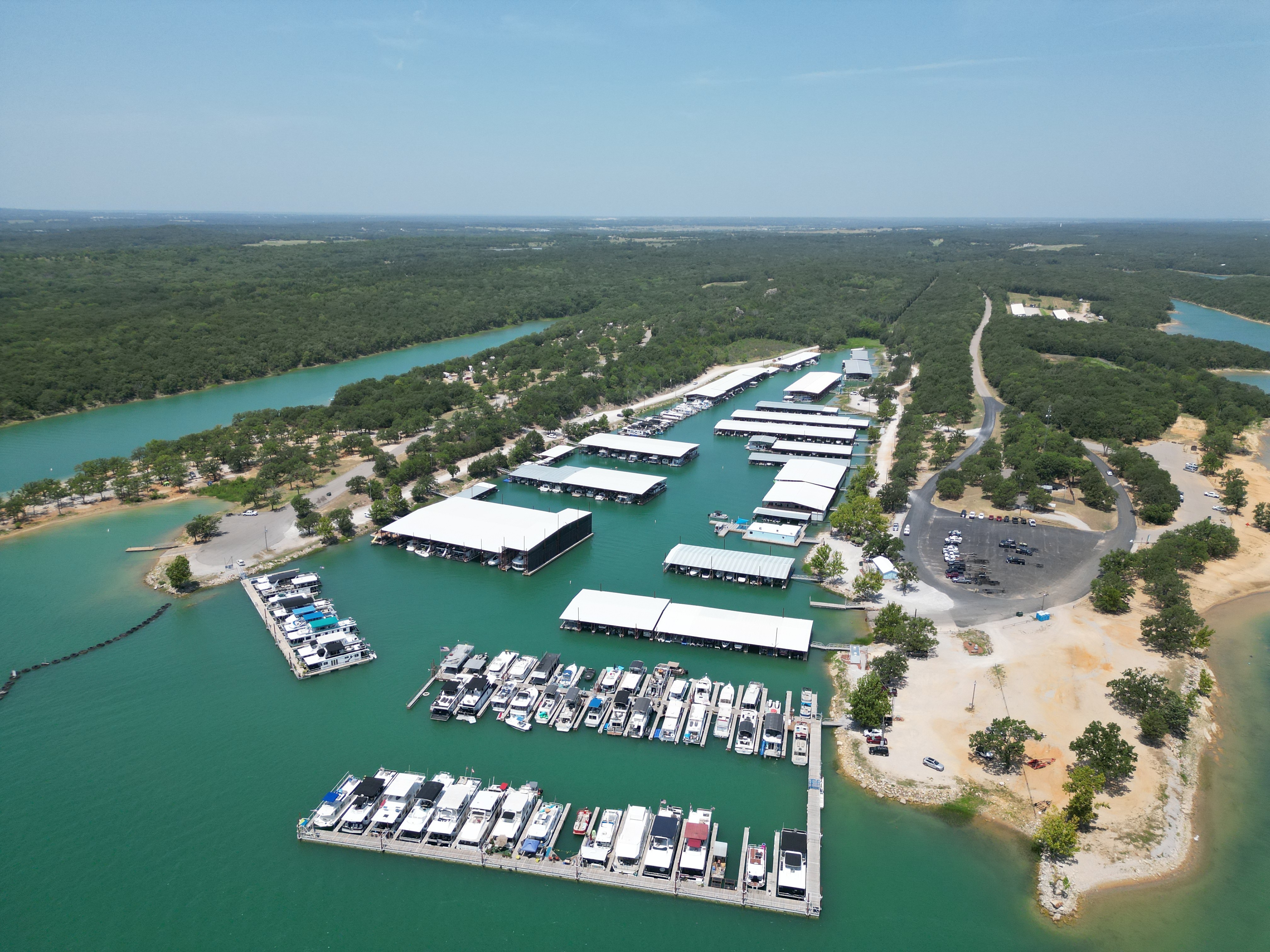 An aerial view of a marina with multiple docks and covered boat slips located in lush surroundings, showcasing a vibrant expanse of turquoise water and forested landscapes.