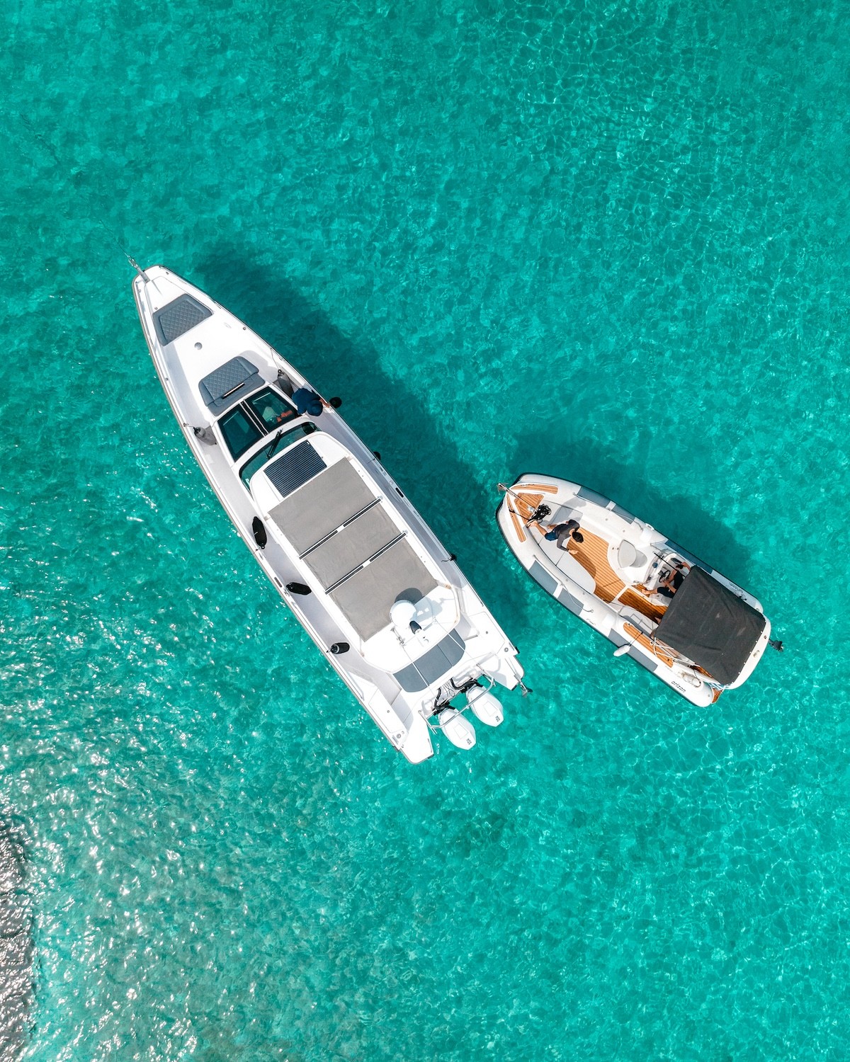 Aerial view of two luxury yachts anchored in crystal-clear turquoise waters of the Cyclades. The larger white motor yacht with twin outboards floats alongside a smaller speedboat with wooden deck details.