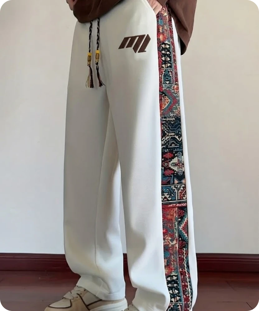 White track pants with a woven carpet-pattern side stripe and the slanted "M" printed in brown at the hip.