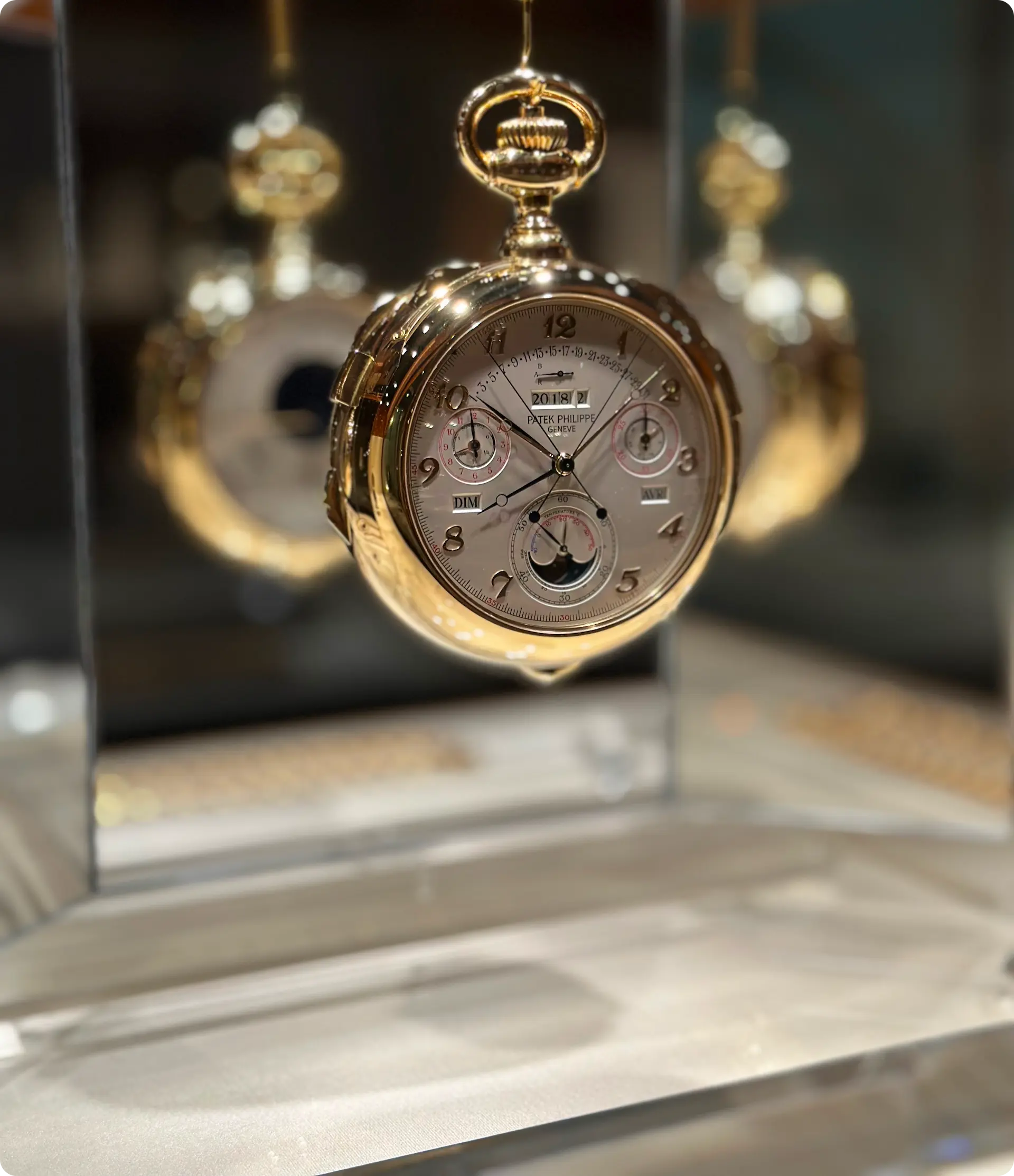 A close-up of a luxurious gold pocket watch with intricate details, displayed in a glass case. The watch face shows multiple dials, including a date window at 12 o'clock, a day window at 9 o'clock, and a moon phase indicator at 6 o'clock. The brand name "Patek Philippe" is visible on the dial.