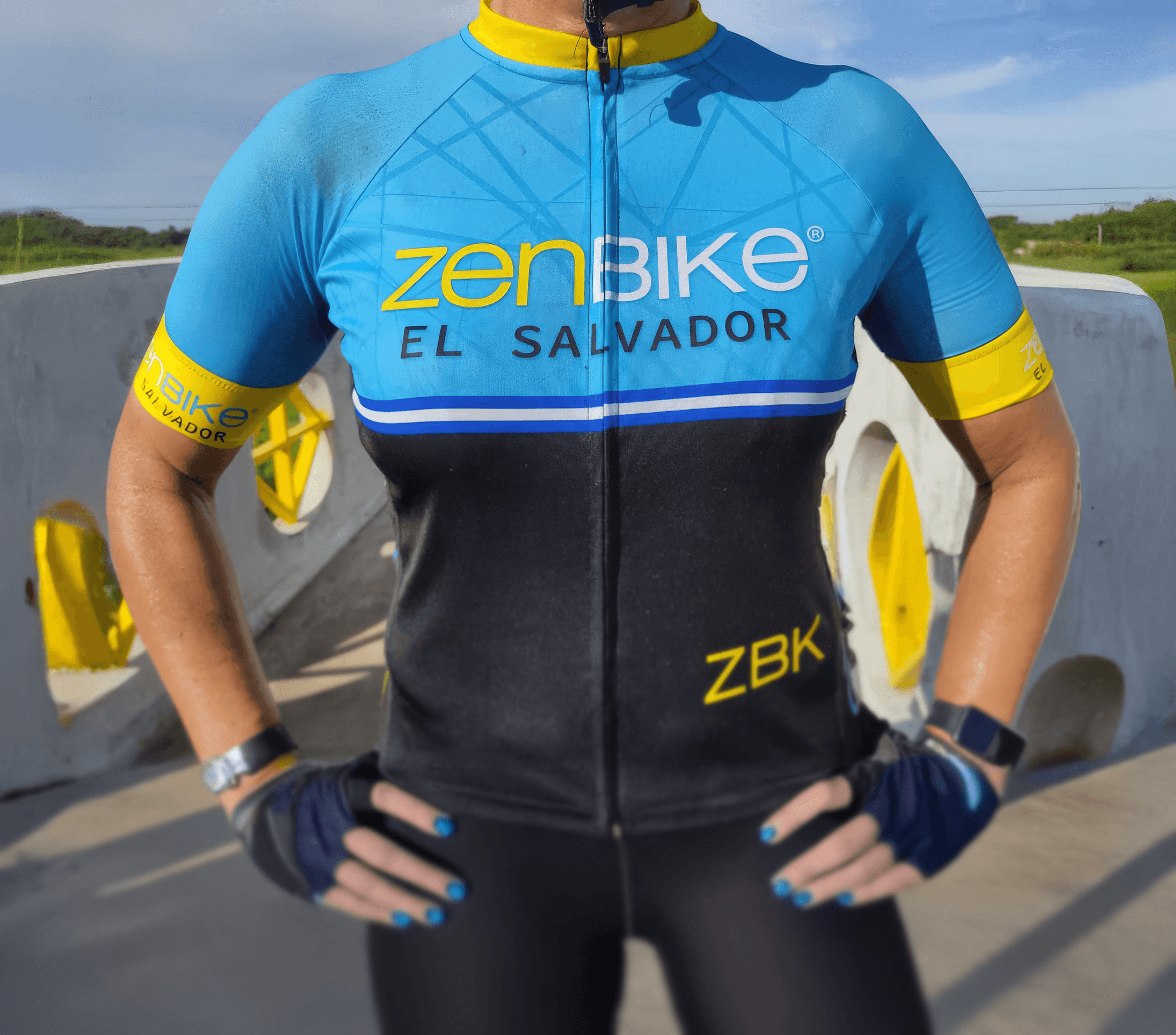 Ciclista con uniforme de zenbike El Salvador posando con manos en la cintura, bajo luz natural y fondo arquitectónico.