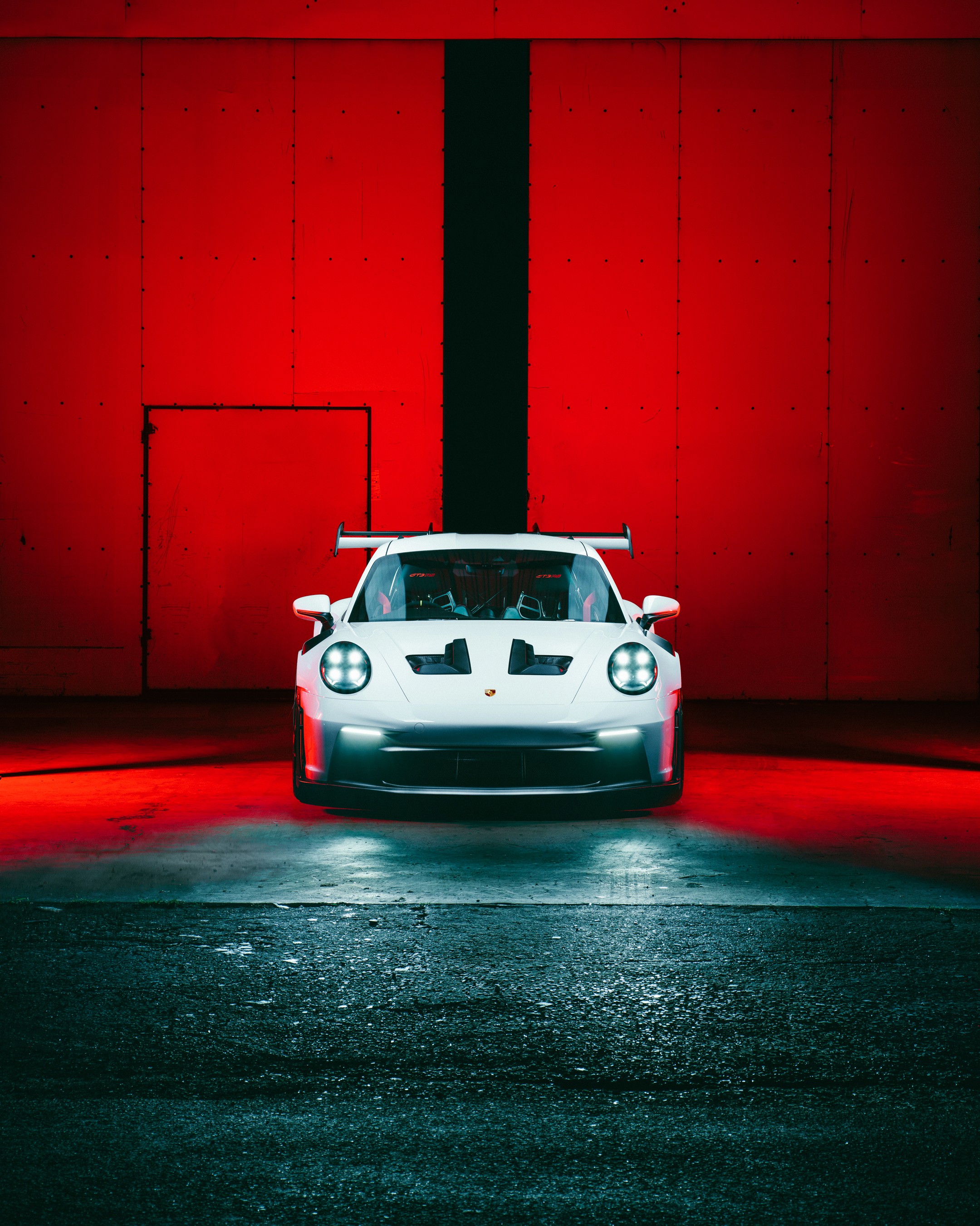 White Porsche 911 GT3 RS (992) shot head-on with all headlights lit, centred against a red industrial roller door, swan-neck rear wing rising above the roofline and bonnet vents in frame, the floor washed in red and cool light, in an industrial setting in Nicosia.