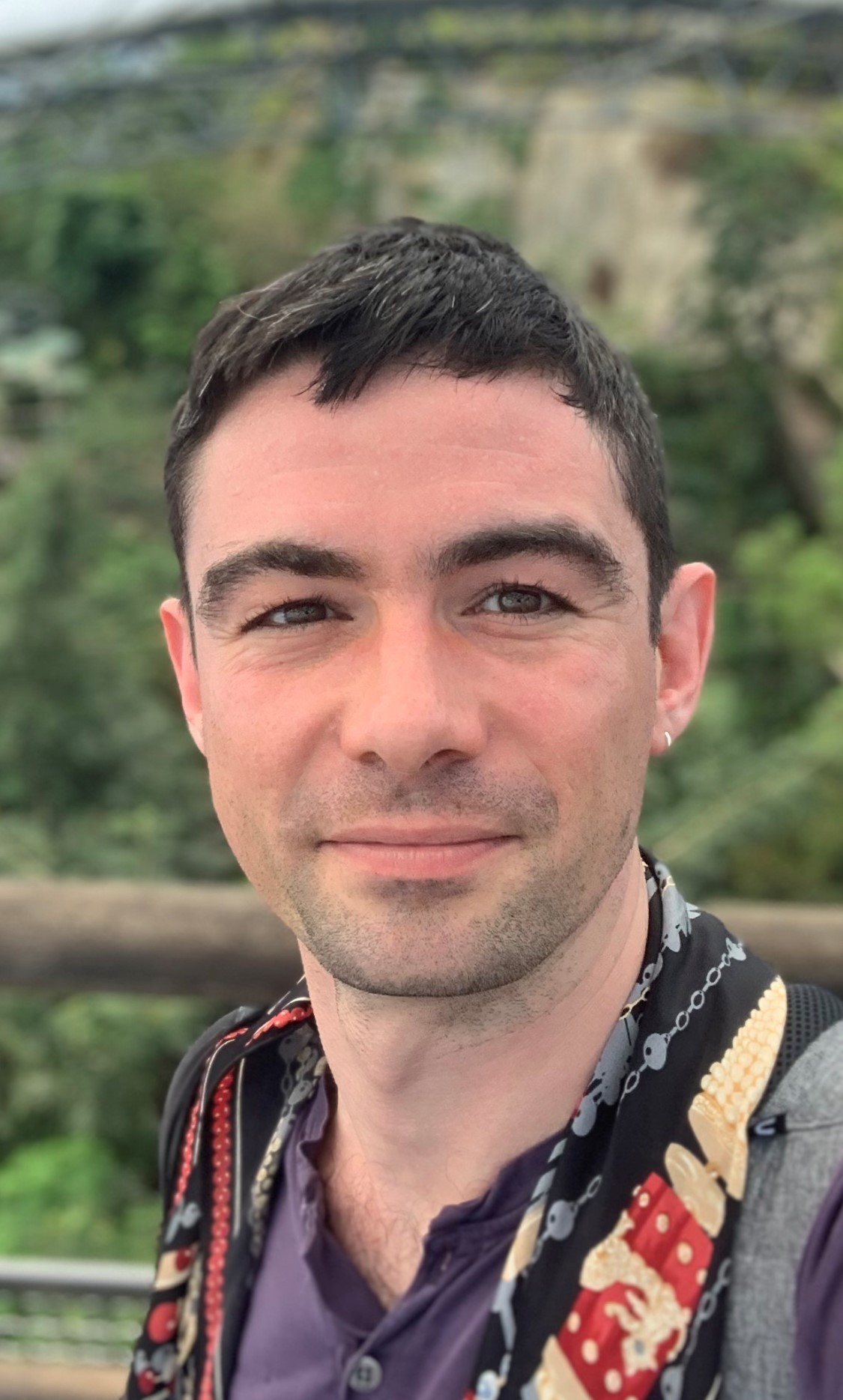 A man smiling wearing a grey jacket and purple shirt.