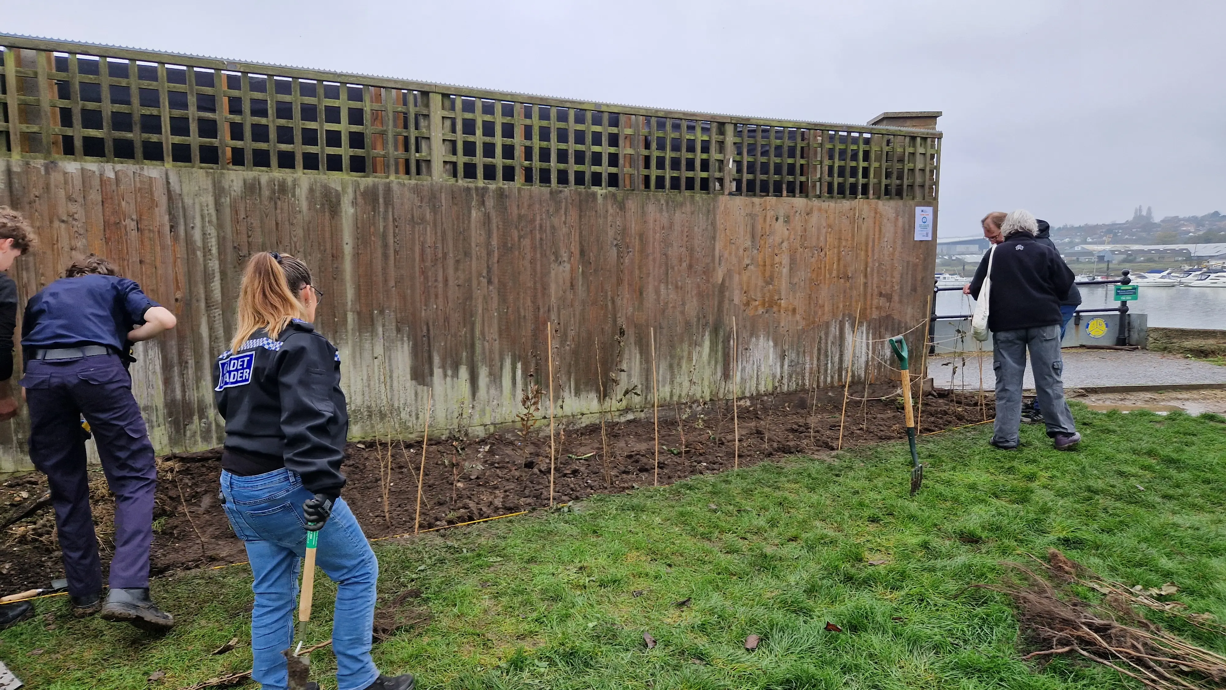 Two people are working near a concrete wall, with another person standing further away on grass, under a cloudy sky.