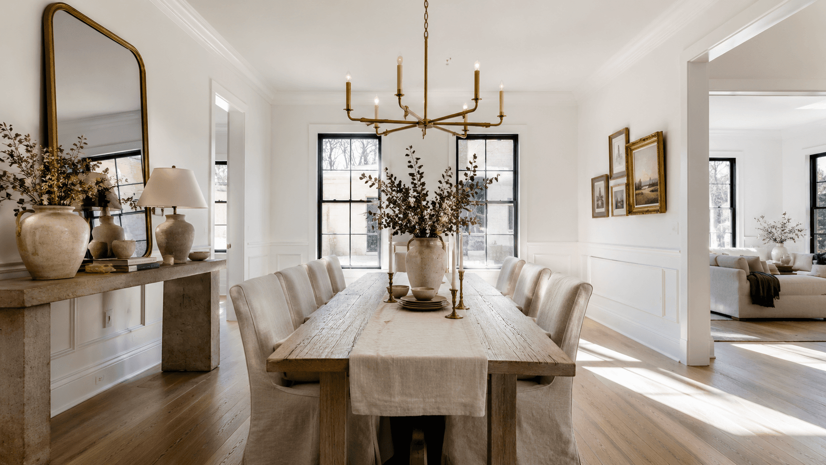 Bright modern French country dining room featuring a long rustic wood dining table, linen slipcovered chairs, brass chandelier, limestone console table with decorative pottery, oversized arched mirror, white paneled walls, and black-framed windows.