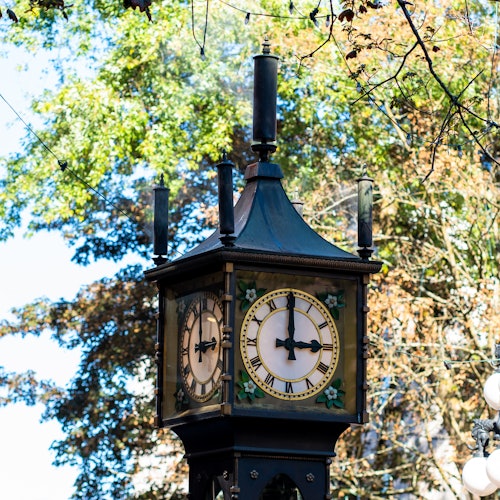 A steam clock with a decorative design, showing two clock faces, surrounded by trees with autumn leaves.