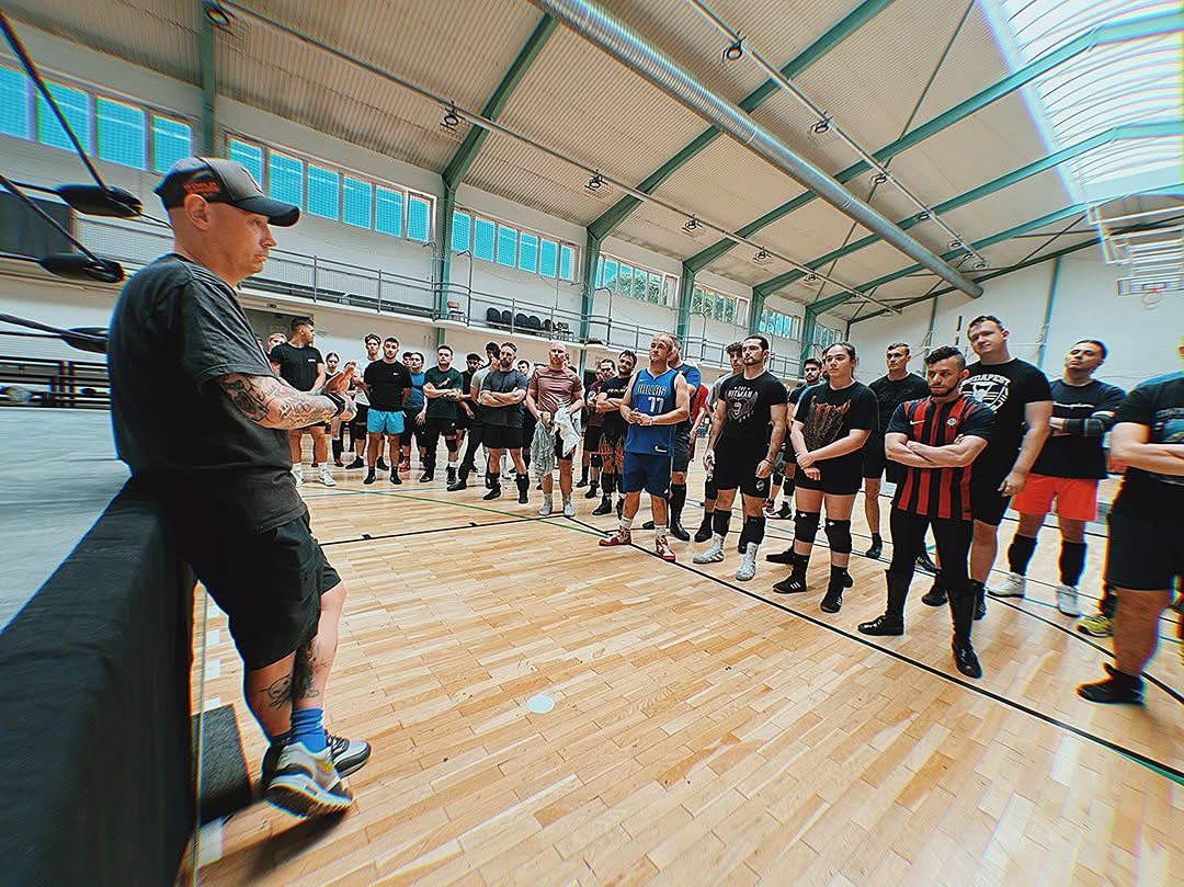 A group of five people dressed in casual, neutral-toned clothing sit around a rustic wooden table in a cozy, stone-walled kitchen, smiling and engaging in conversation with bowls and a covered dough resting in front of them, creating a warm, communal atmosphere.