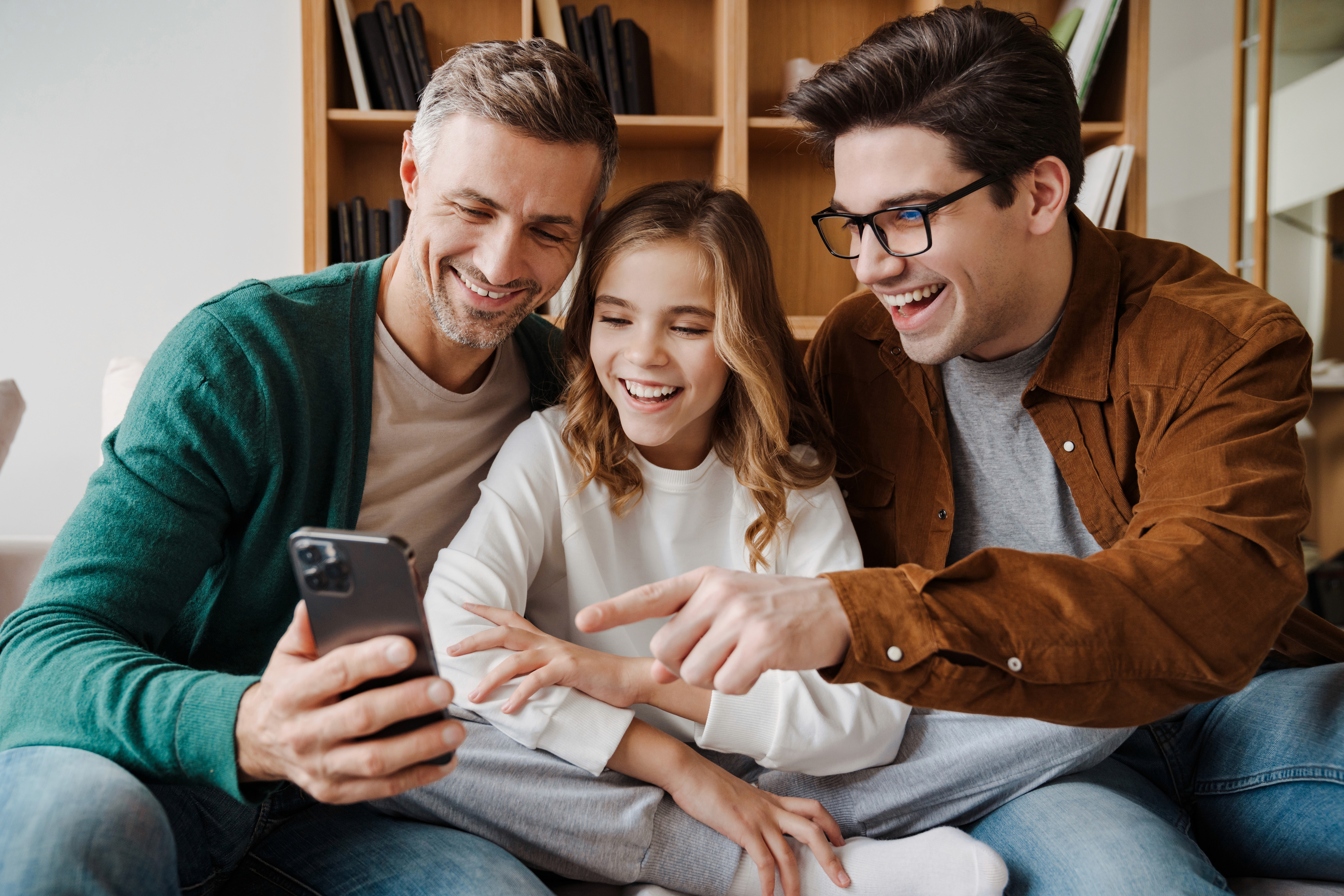 Zwei Väter und ihre Tochter lachen gemeinsam beim Blick auf ein Smartphone – moderne Regenbogenfamilie genießt gemeinsame Zeit zu Hause.