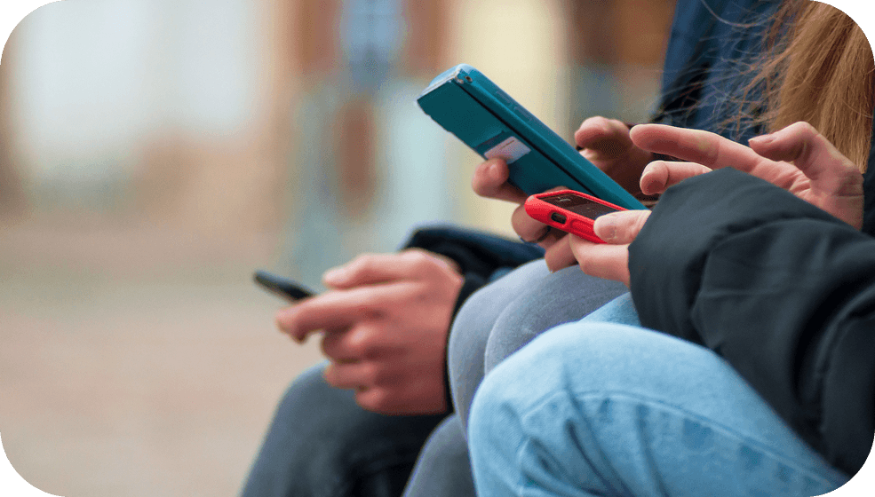Close-up of teenagers sitting side by side outdoors, each scrolling on their smartphones, showing everyday teen social media use