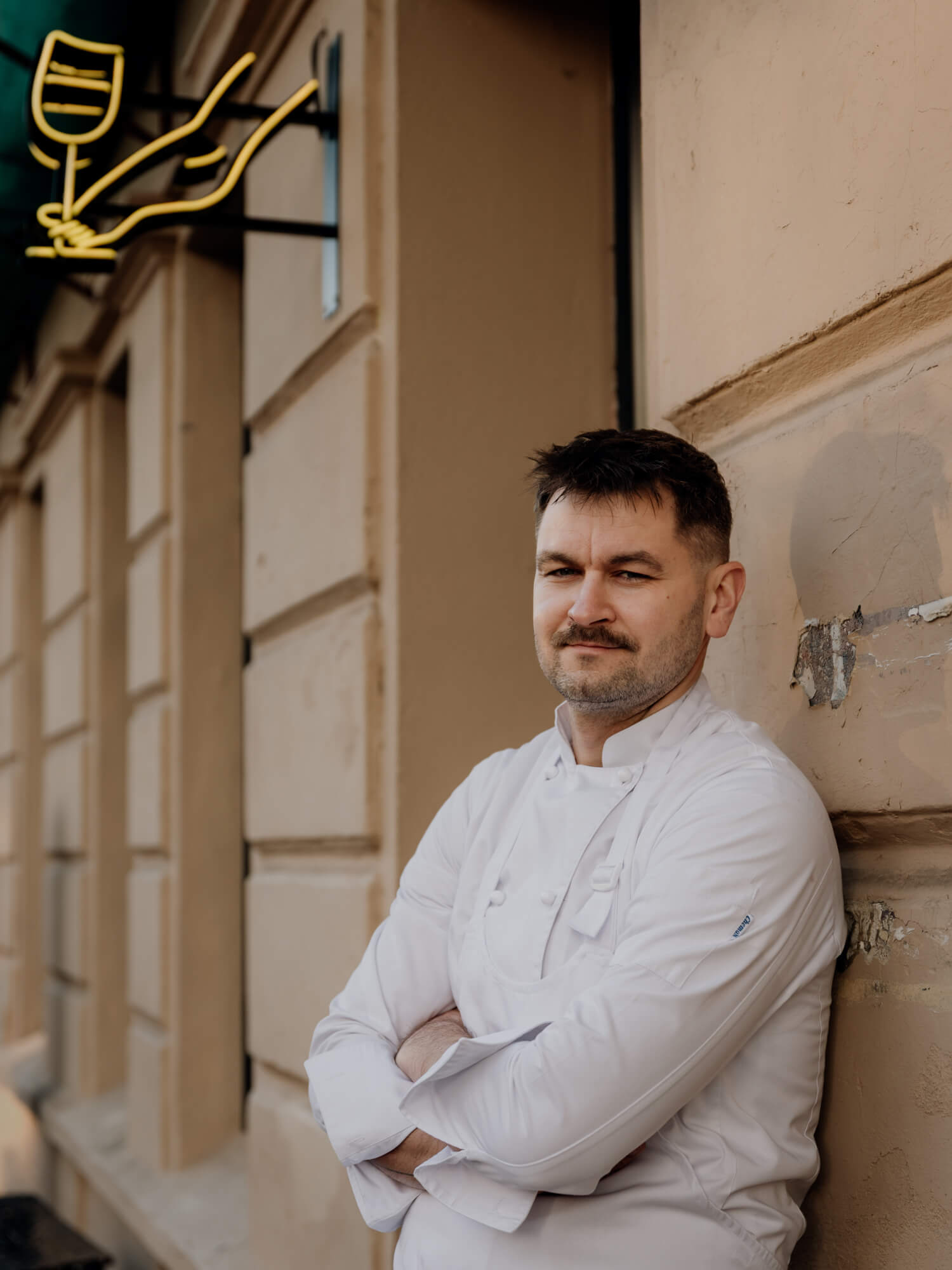 A person stands casually with arms crossed against a building wall, wearing a light sweater and looking thoughtful.