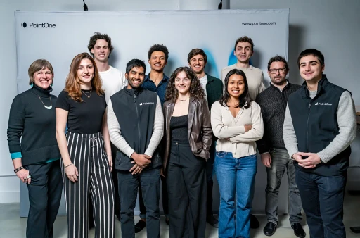 A diverse team of eleven smiling PointOne employees, dressed in smart casual attire, stands together against a branded.