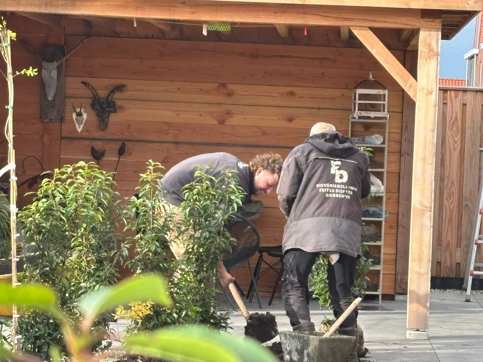 Twee hoveniers planten struiken in een rustige, groene tuin met houten overkapping en terrastegels in De Vrieswijck Gorredijk.