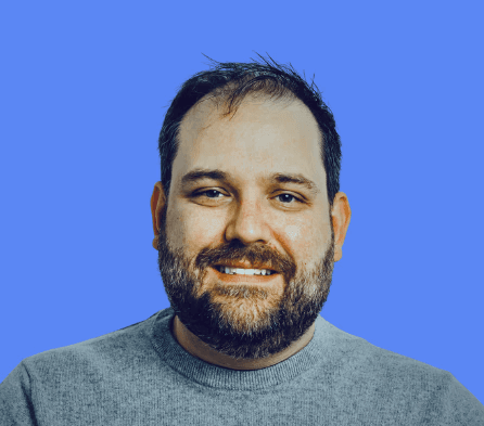 A smiling man with curly hair is sitting at a desk, looking at the camera, with a laptop and a lamp in the background.