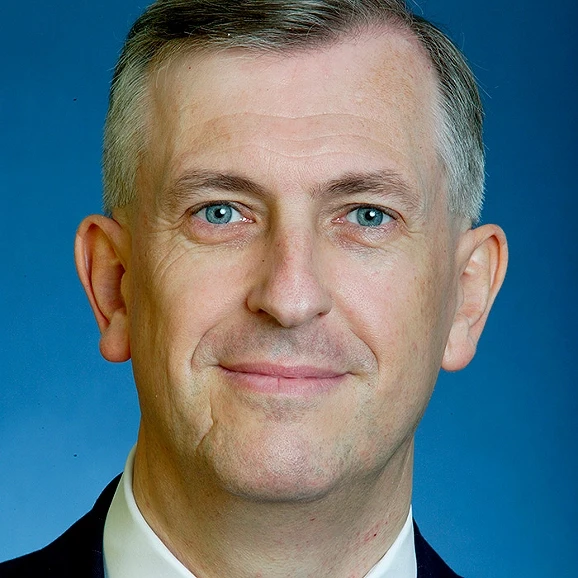 A focused woman with long red hair is seated at a desk, wearing a black blazer and dark blouse. She intently looks at camera, smiling slightly, with an open laptop in front of her. The setting is a warm and serene office setup in the background. Soft daylight filters through, creating a calm atmosphere. The composition captures her in a frontal profile with a clear emphasis on her concentration and the serene indoor setting.