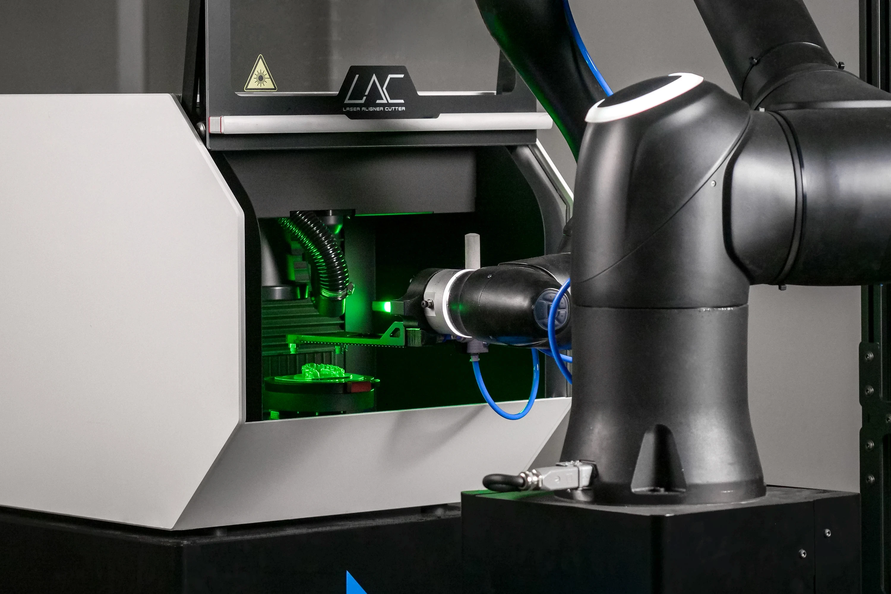 A robotic arm efficiently tending a DMU LAC machine by loading a clear aligner for cutting in a close-up view with a neon green glow illuminating the inside of the machine.