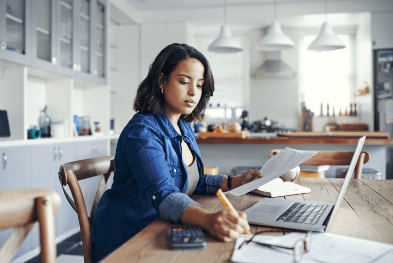 Woman sitting at a kitchen table, writing notes and working on a laptop, representing care home staff scheduling and rota planning