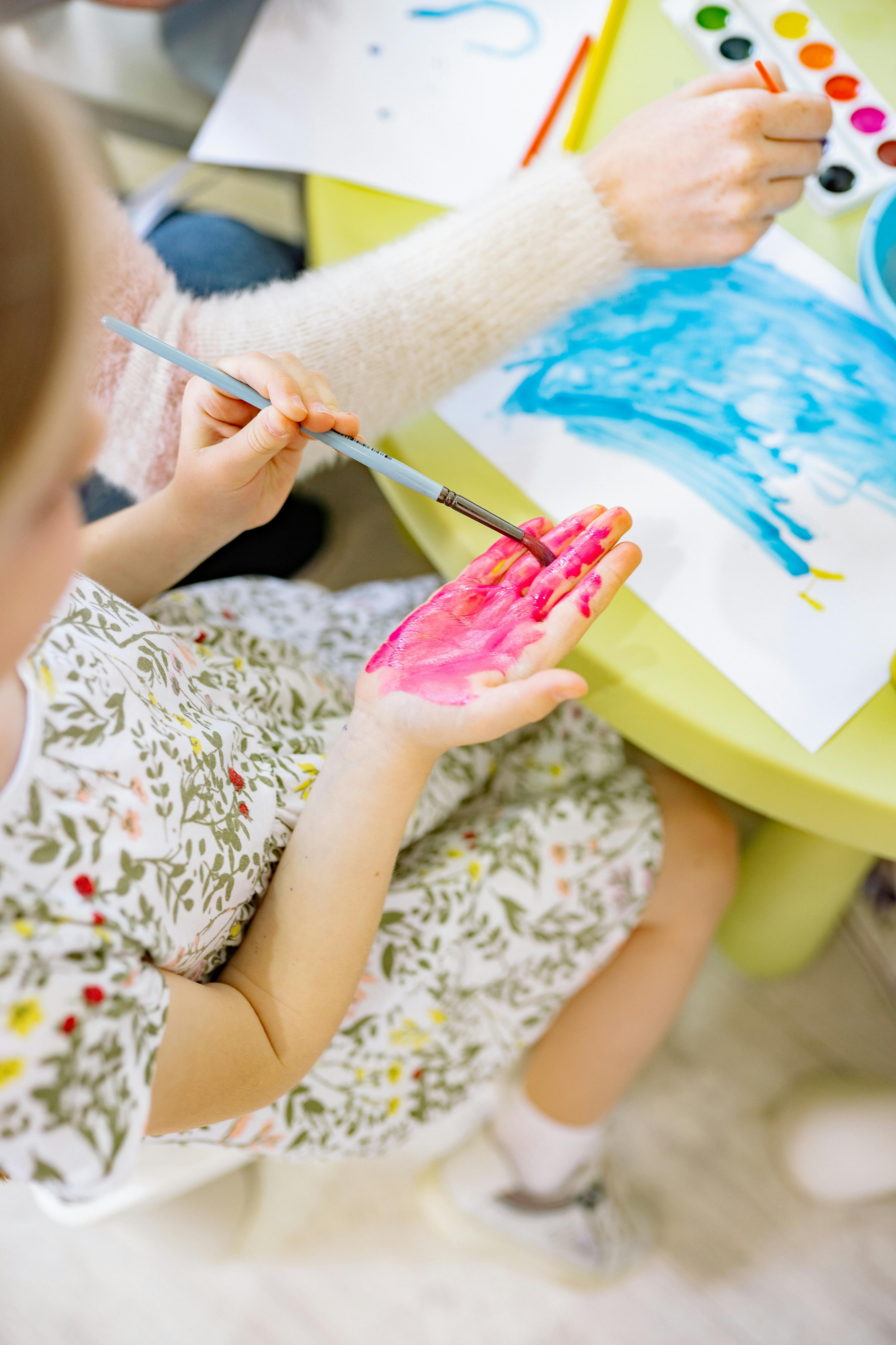Vista superior de un niño o niña con la mano pintada de rosa durante un taller de arteterapia en San Fernando. El niño sostiene un pincel sobre una cartulina con acuarelas azules, acompañado por una terapeuta en Espacio Yutori.