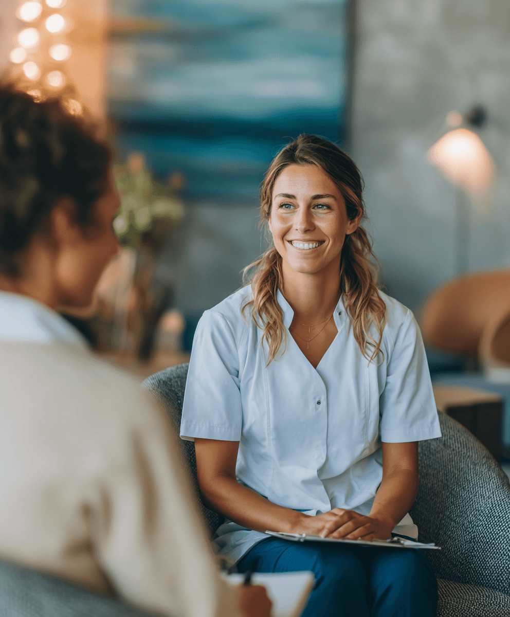 A smiling woman in a white uniform holds a clipboard, sitting in a cozy, warmly lit room. She appears engaged, suggesting a friendly consultation.
