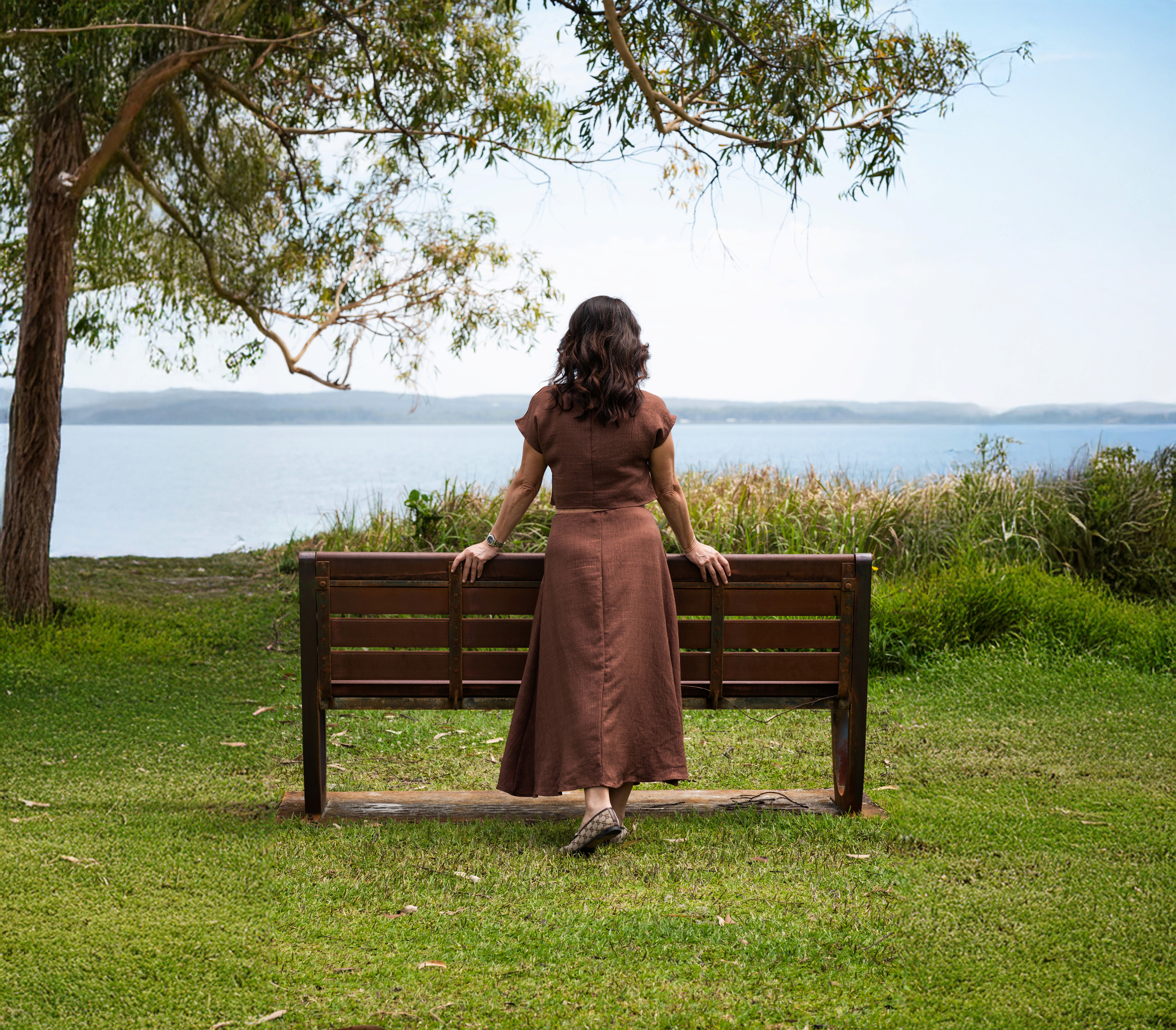 A woman in a brown dress sits on a bench by a grassy area near the water, under a tree.