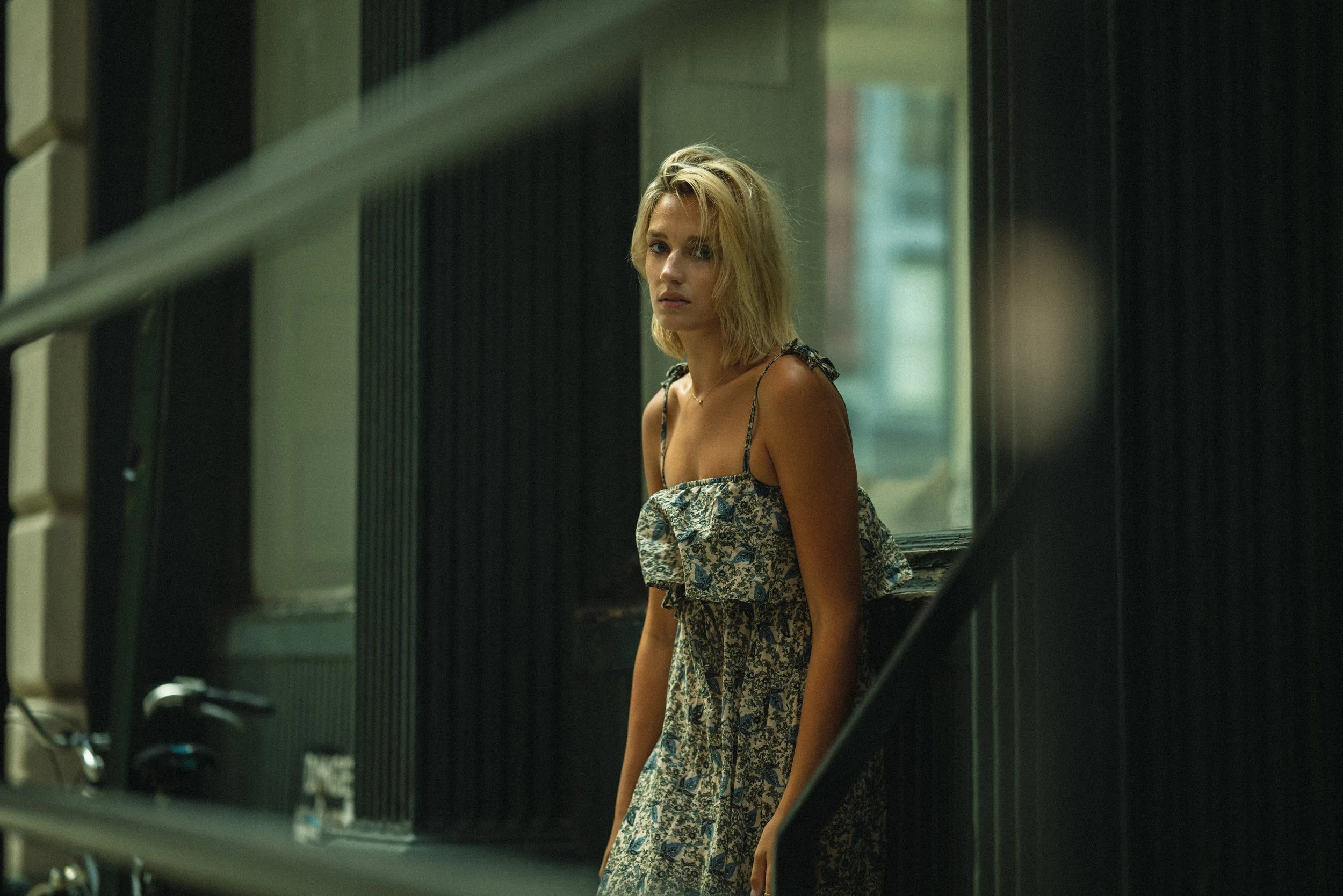 Blonde woman in a blue floral sundress in a dim industrial loft, glancing over her shoulder toward a sunlit window.
