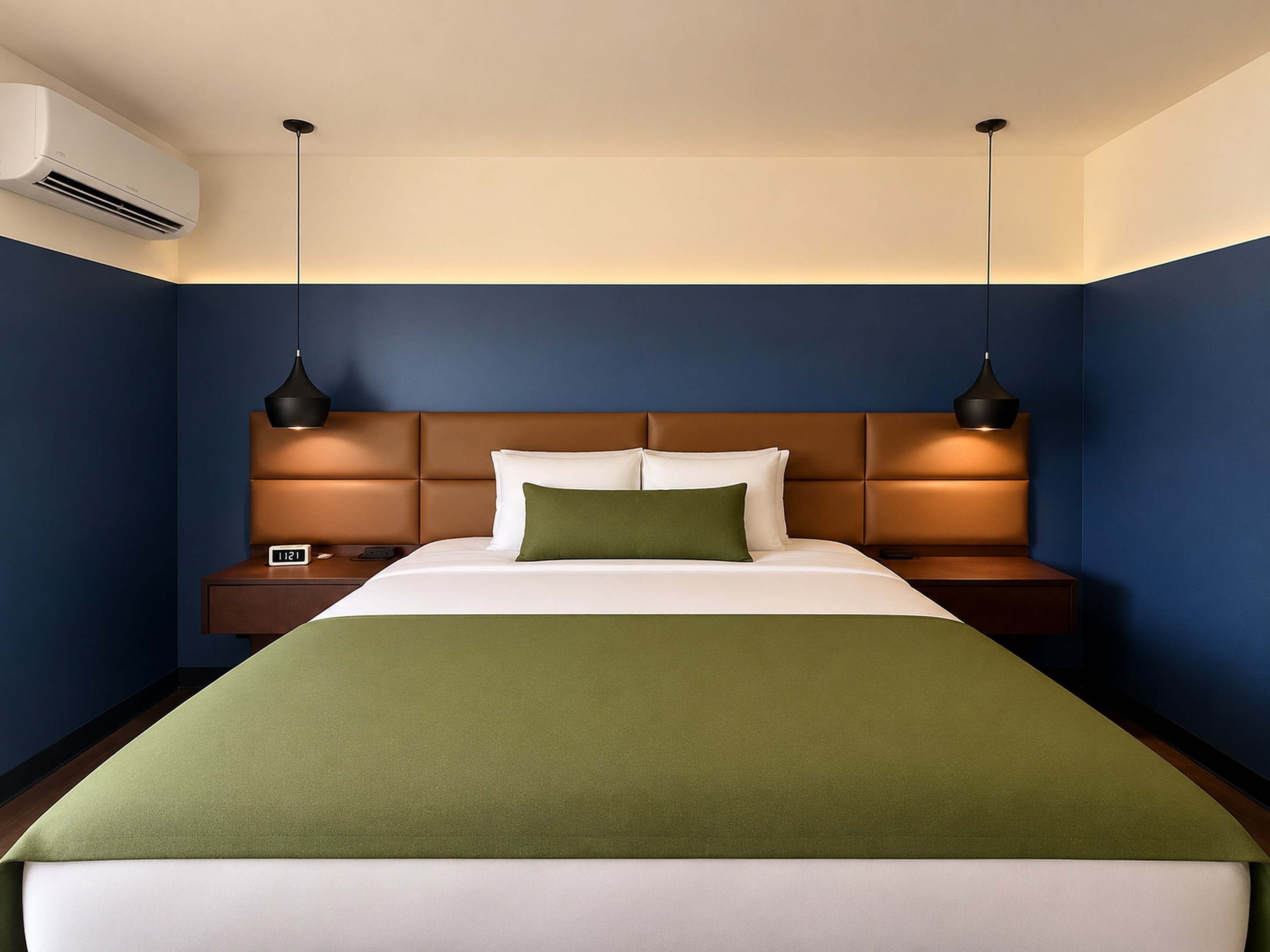 A renovated Superior King room at The Wesley, with a king bed, tan leather headboard, deep blue walls, olive green blanket and bolster, and two black pendant lights.