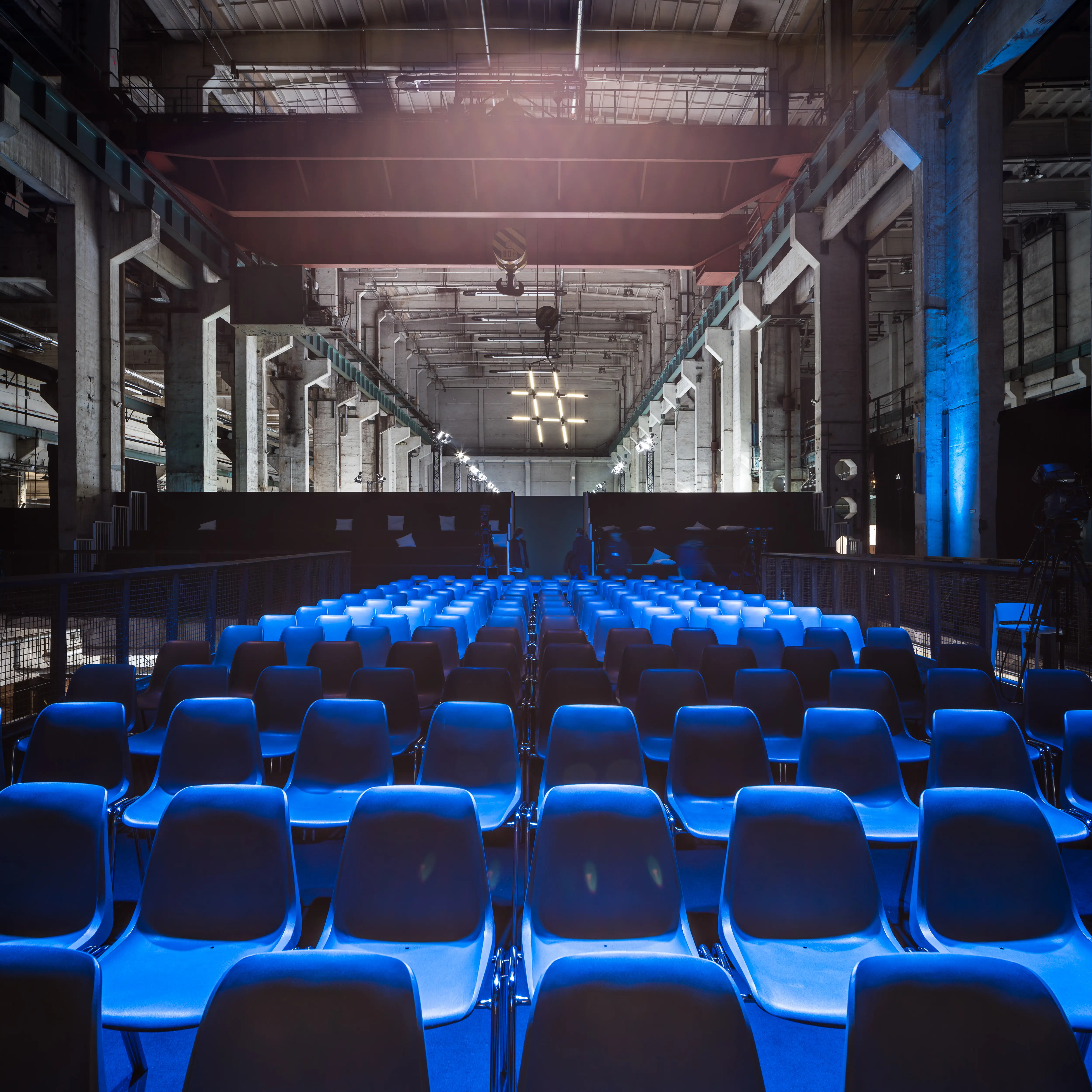 Blue shell chairs in rows before the NEONYT conference stage, backlit by daylight at the end of the monumental hall.