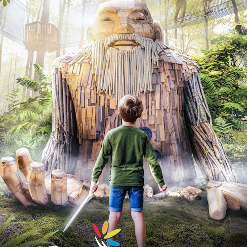 一个男孩手持玩具剑站在一个郁郁葱葱的室内花园里,花园中有蕨类植物和树木,还有一个大型木制人形雕塑。