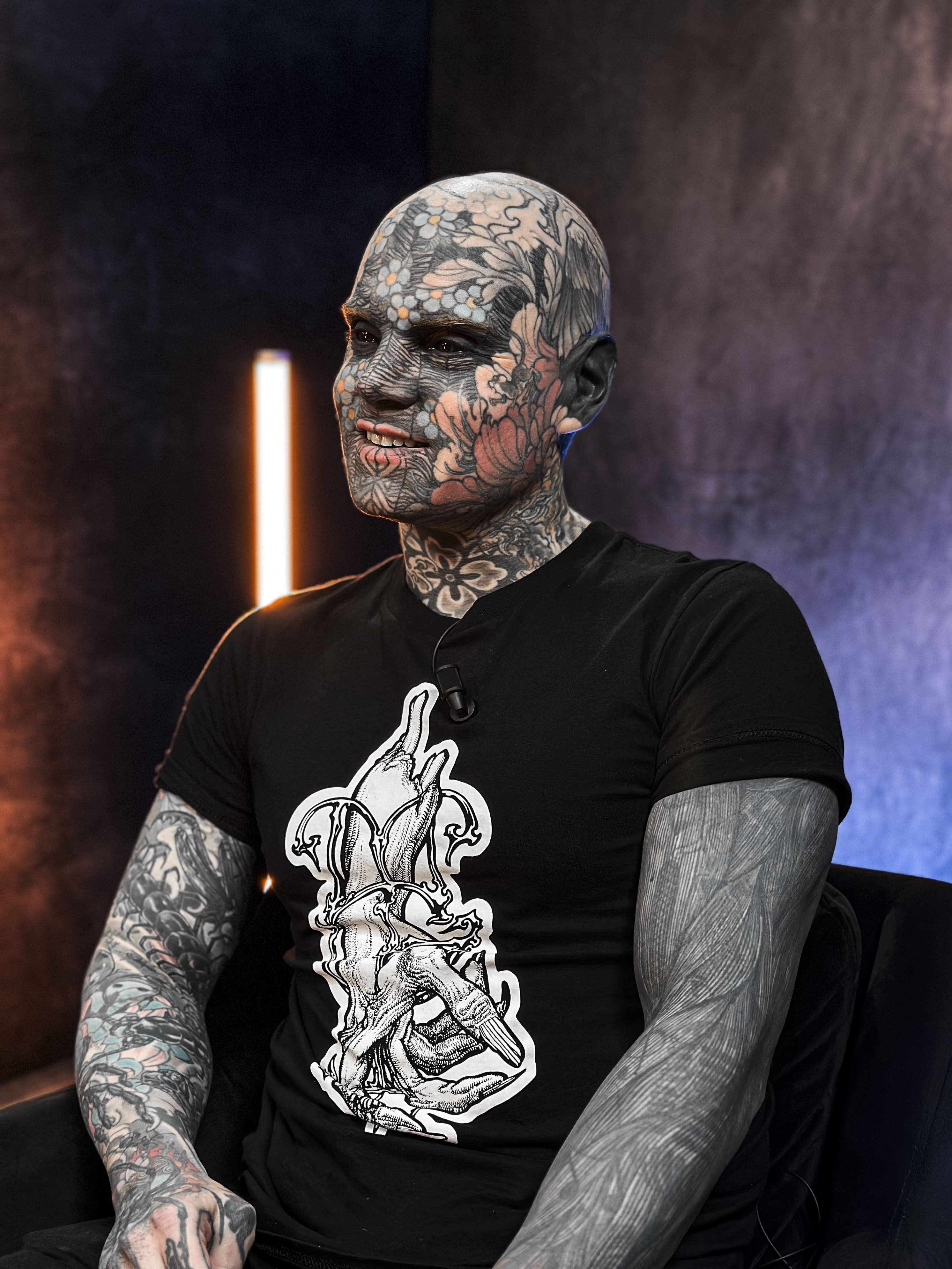 Photographie de portrait professionnelle d'un homme au corps et au visage entièrement tatoués, y compris le blanc des yeux (tattooed blackwork). L'invité est assis, portant un T-shirt noir avec un graphisme stylisé. Il sourit légèrement, regardant vers la gauche. L'éclairage de studio utilise un fond sombre avec des néons verticaux (orange et bleu/violet) créant une ambiance dramatique et artistique. Cette photo de personnalité, retouchée par Nux-Studio, est un exemple de production photo pour des interviews sur des sujets de société, des cultures alternatives ou le body art.