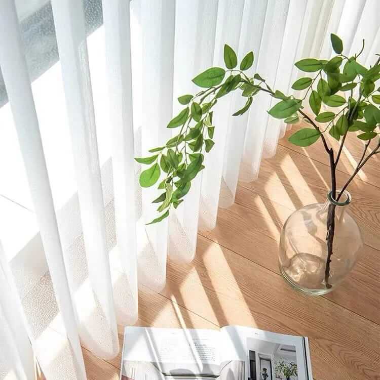 Sunlight filters through sheer shades onto a wooden floor, illuminating a glass vase with green leaves and an open magazine.