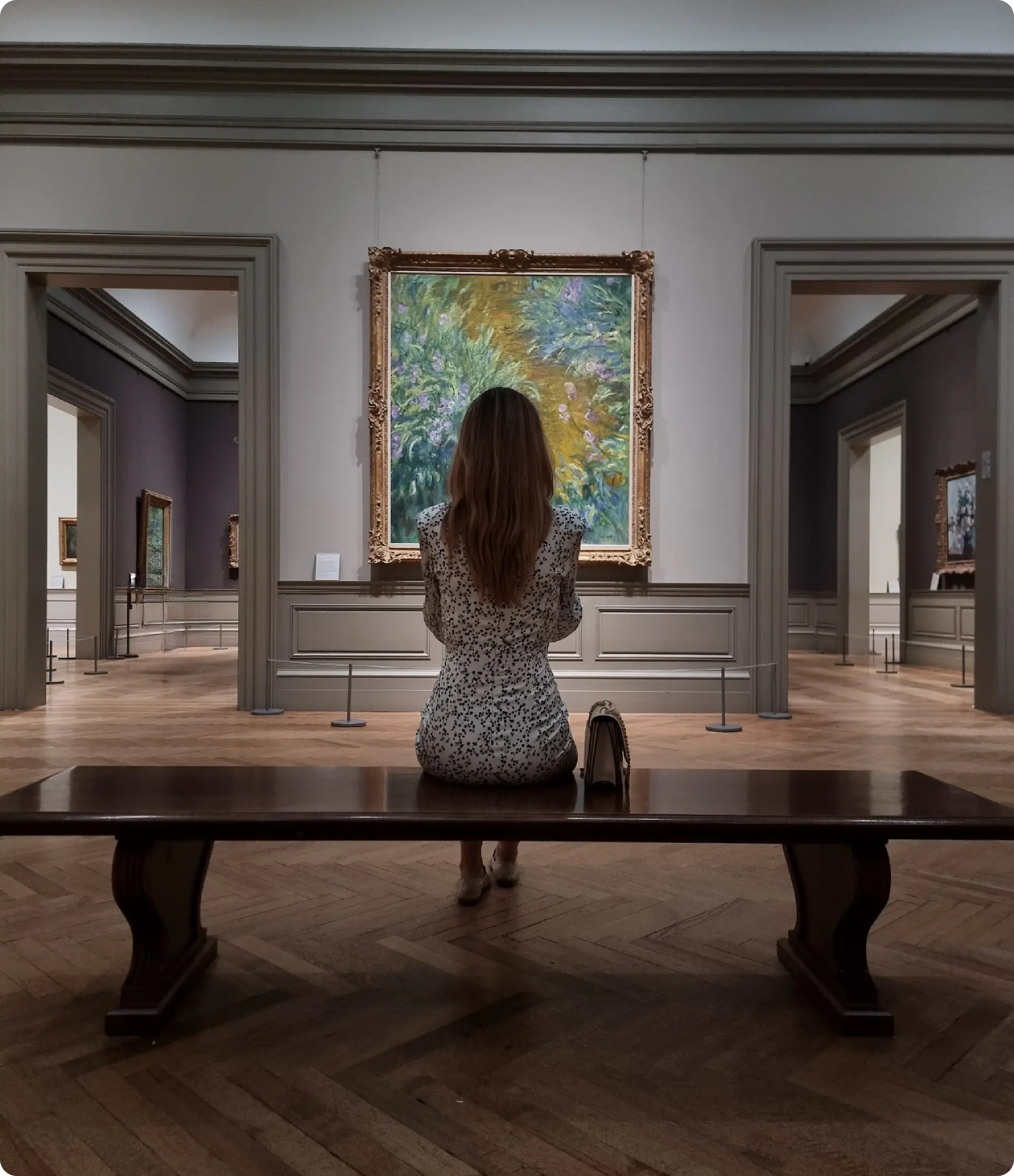 A woman with long hair, wearing a white dress with a black pattern, sits on a wooden bench in an art gallery. They are facing a large, framed painting on the wall, which features abstract, colorful brushstrokes. The gallery has wooden floors, gray walls, and other artworks visible in the background.