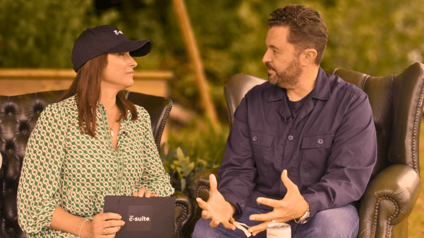 Young person with short, tousled blonde hair wearing an oversized, vibrant orange turtleneck sweater against a soft grey background, conveying a cool, modern look.