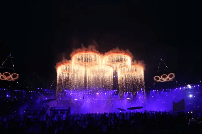 A stunning display features illuminated Olympic rings with cascading pyrotechnics, lighting up the night sky amid a vibrant, cheering crowd at a grand stadium event.