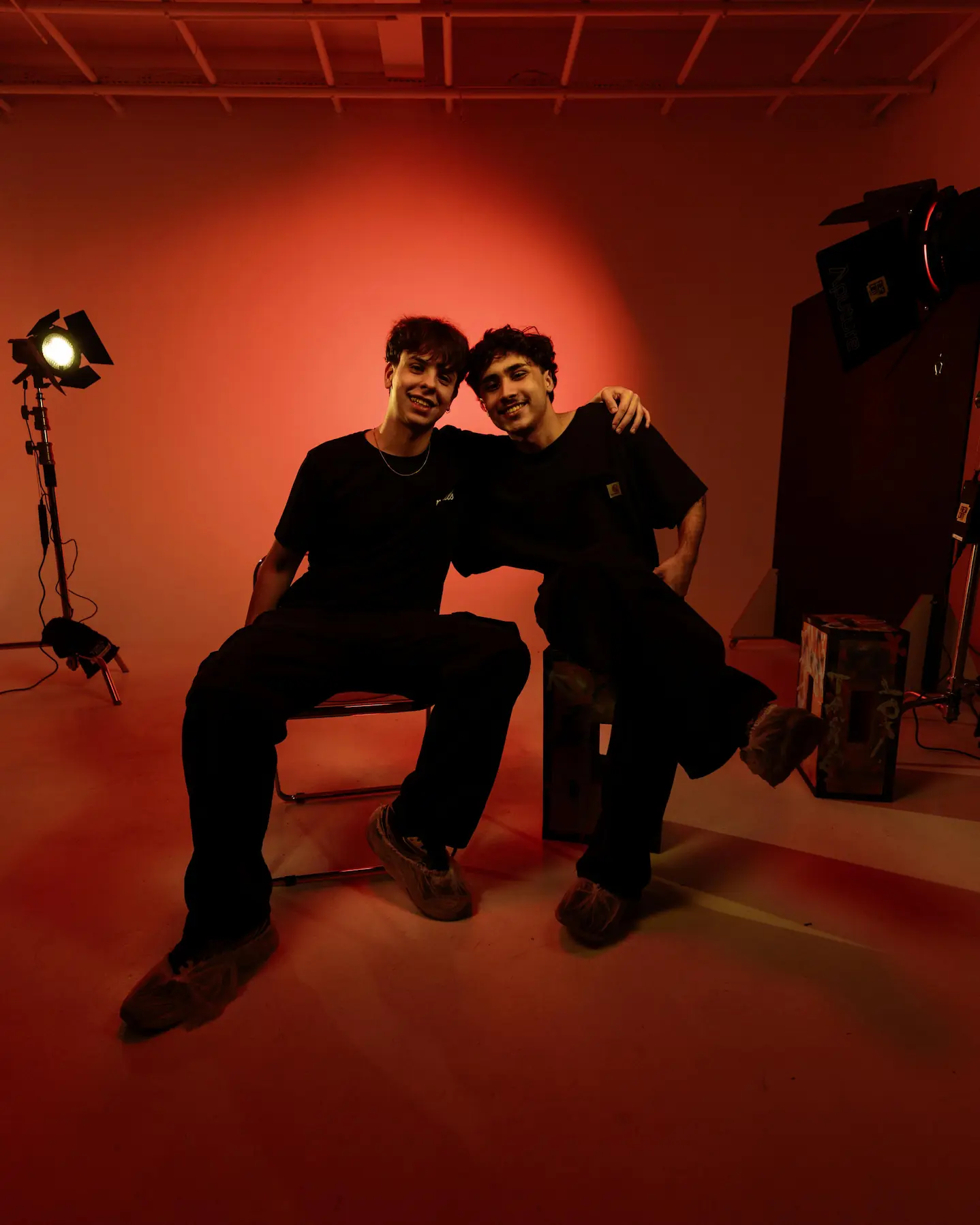 Two men in black smile under red studio lighting, arms around each other, surrounded by professional equipment.