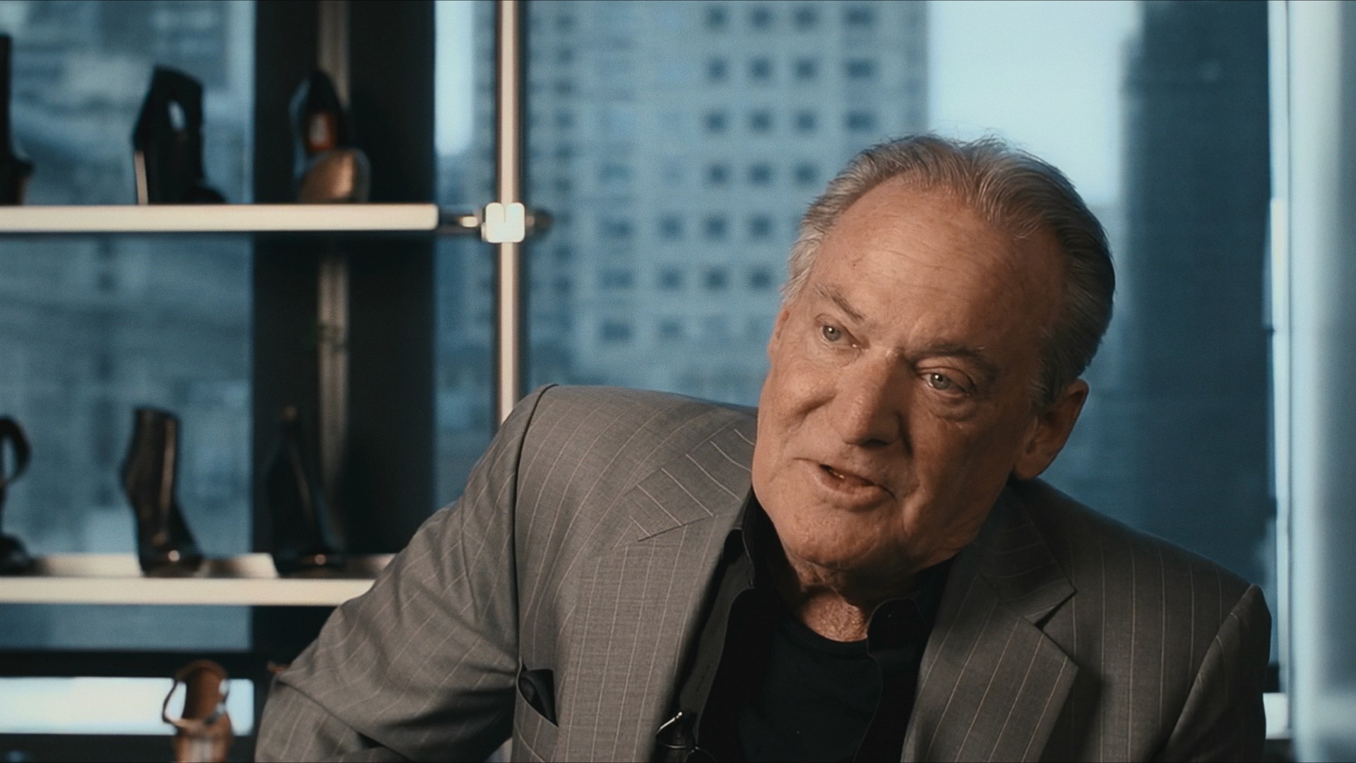 A man in a grey jacket interviewed beside a shelf of heels, the city behind him, Maddman