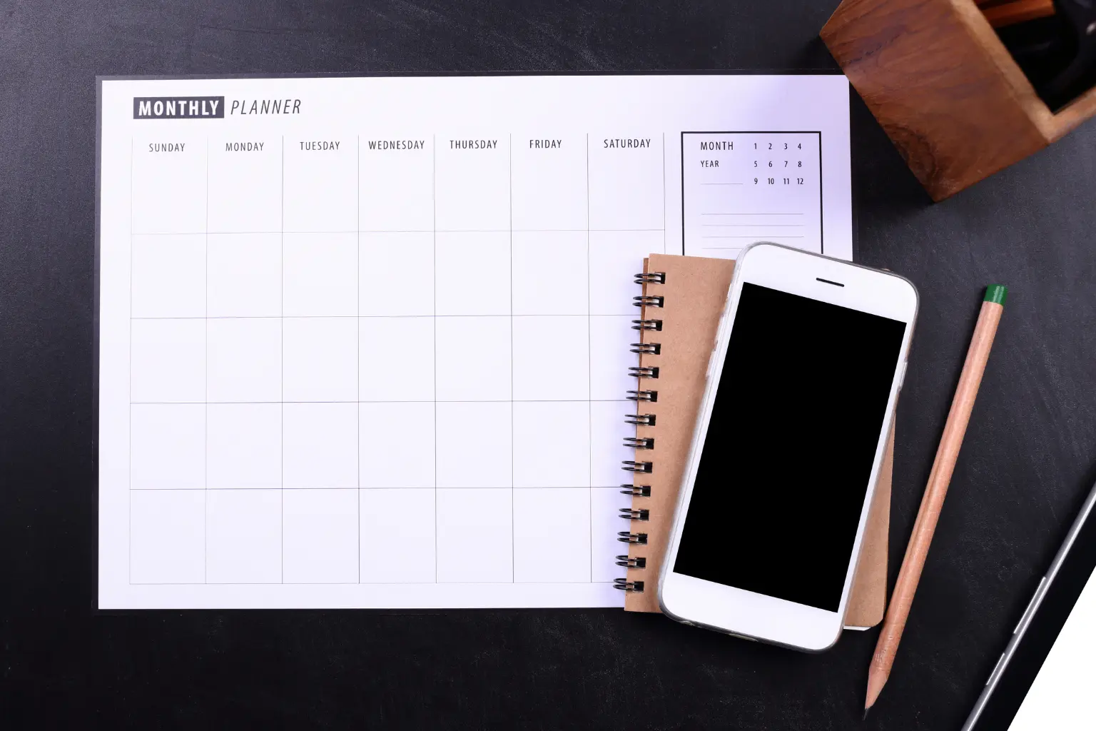 Automated calendar layout featuring a blank monthly planner beside a smartphone, pencil, and notebook on a black desk