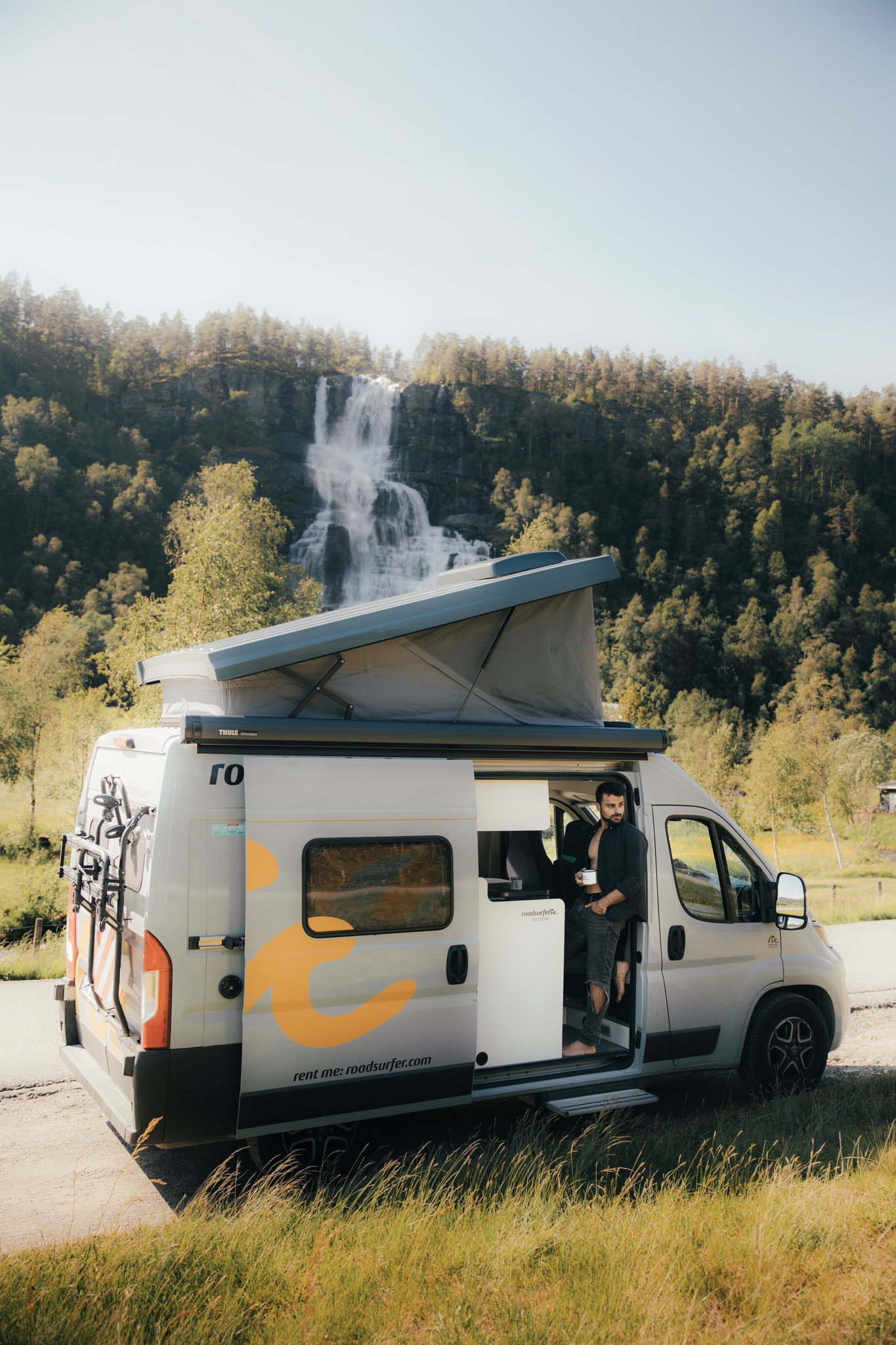 A camper van parked beside a road, with its roof extended, set against a backdrop of trees and blue sky.
