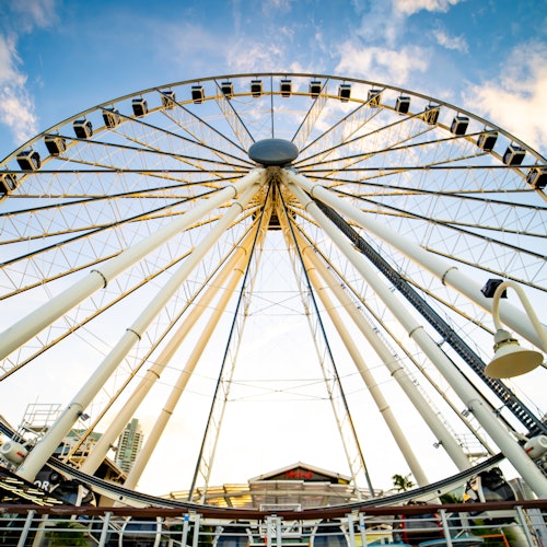 Vista en ángulo bajo de una gran rueda de la fortuna contra un cielo azul con nubes dispersas. La base de la rueda tiene un edificio pequeño.