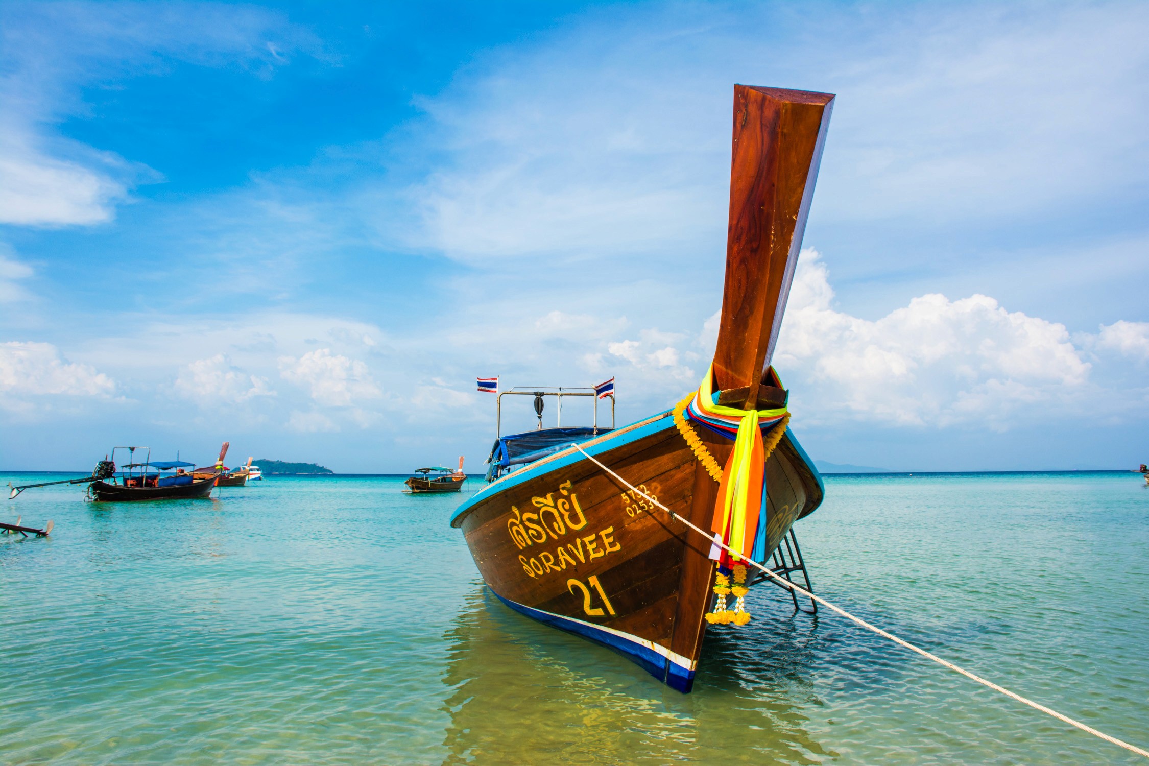 A traditional wooden longtail boat with a colorful prow sits in turquoise water under a bright blue sky and white clouds.