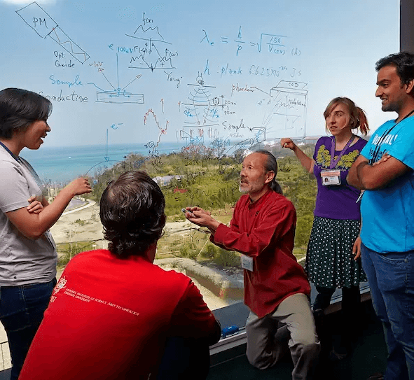 A small group of people, including a professor and students, discussing scientific formulas in front of a whiteboard with a scenic ocean view behind them.